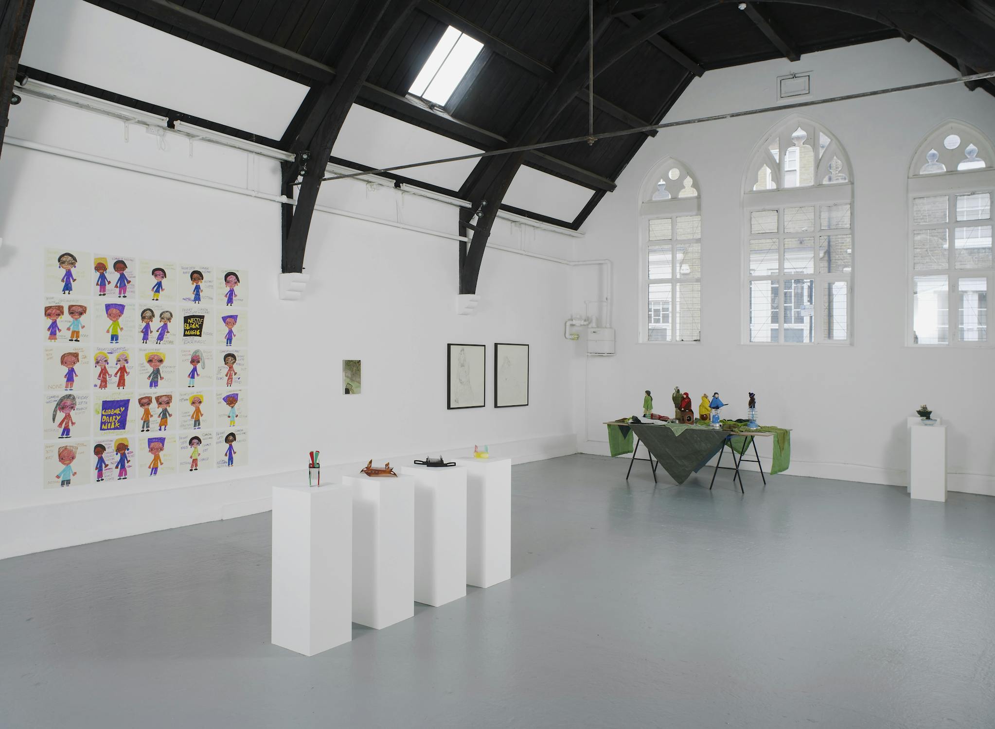 A bright, modern art gallery with white walls, tall arched windows, and a dark wooden ceiling. Artworks, sculptures on pedestals, and a table with colourful objects are displayed throughout the space.