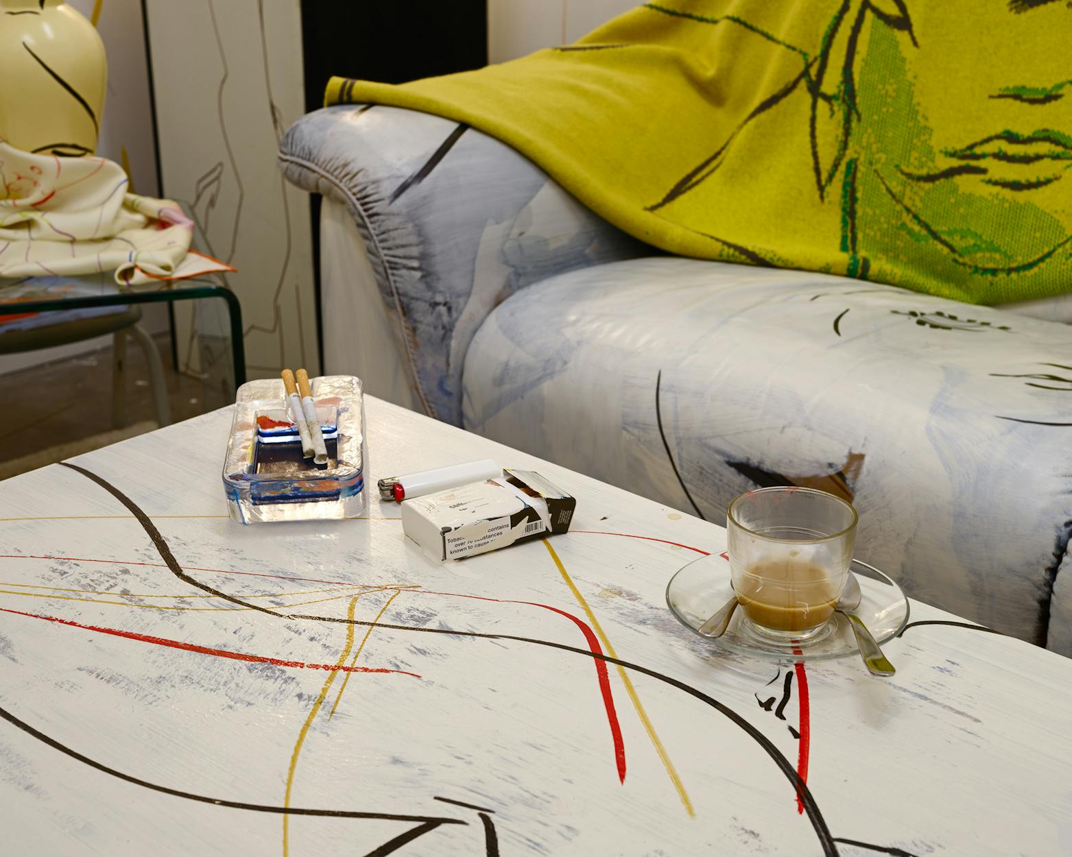 A painted coffee table with abstract lines holds a glass cup with coffee, a saucer and spoon, a carton of cigarettes, an ashtray with cigarette butts, and matches; a couch with a green and yellow blanket is in the background.