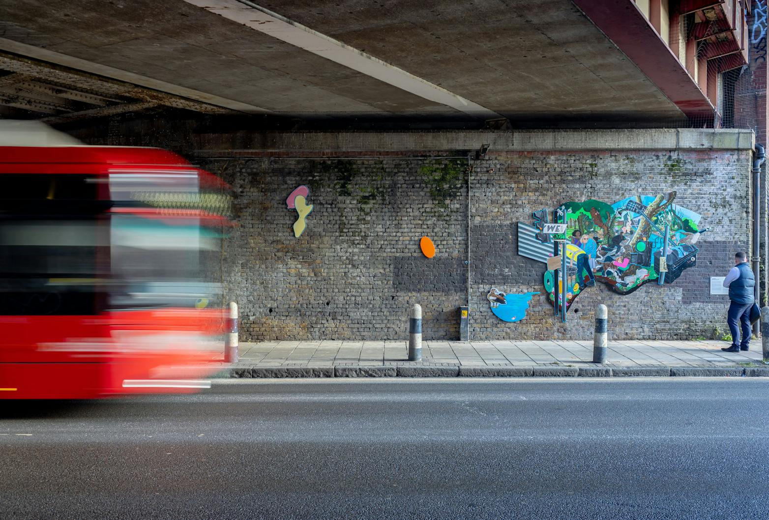 A vibrant and eclectic mural by Flo Brooks is painted on a brick wall. The mural features various elements like a man holding a baby, wildlife such as birds, and urban imagery including street signs and graffiti. There's also a small plaque with text on the wall beside the mural.
