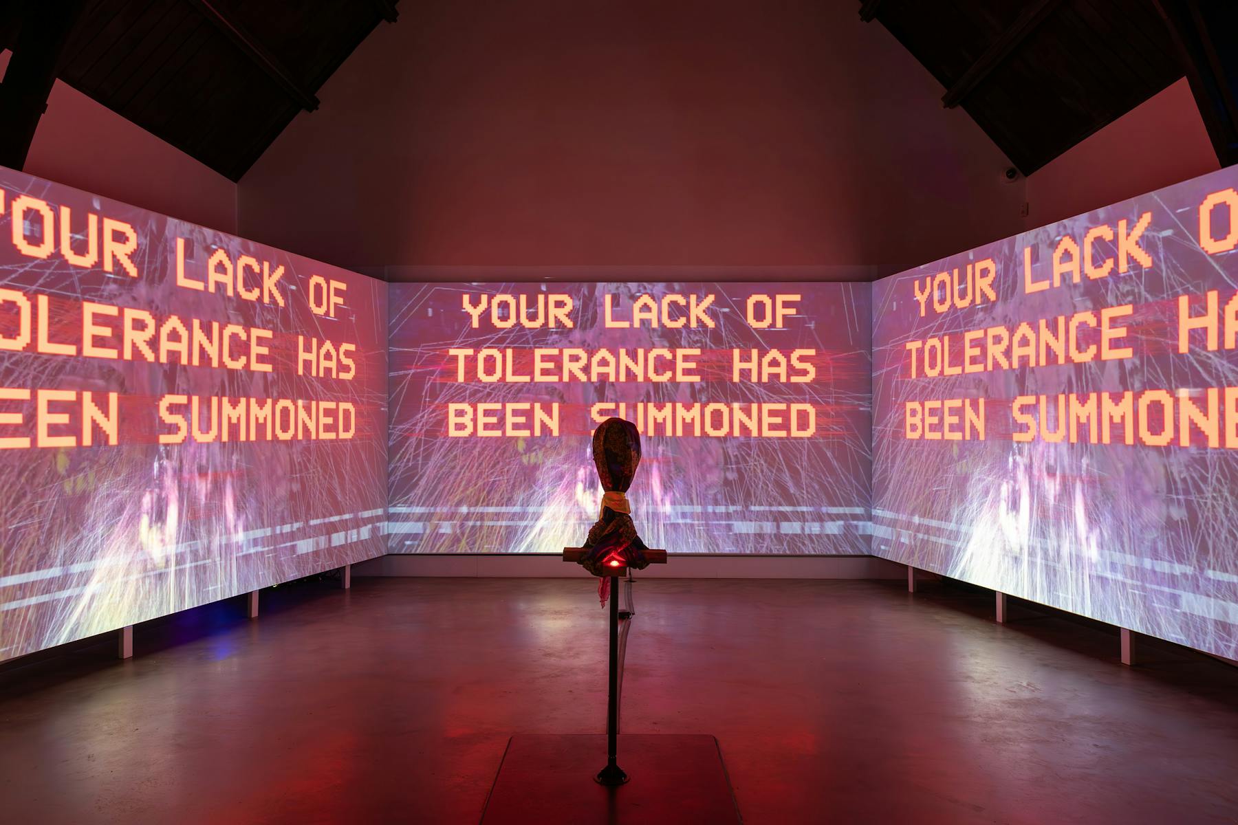 a photograph of three screens that are projected with bright and graphic images. There is text that reads 'YOUR LACK OF TOLERANCE HAS BEEN SUMMONED'. In the foreground there is a hooded object on a stand. 