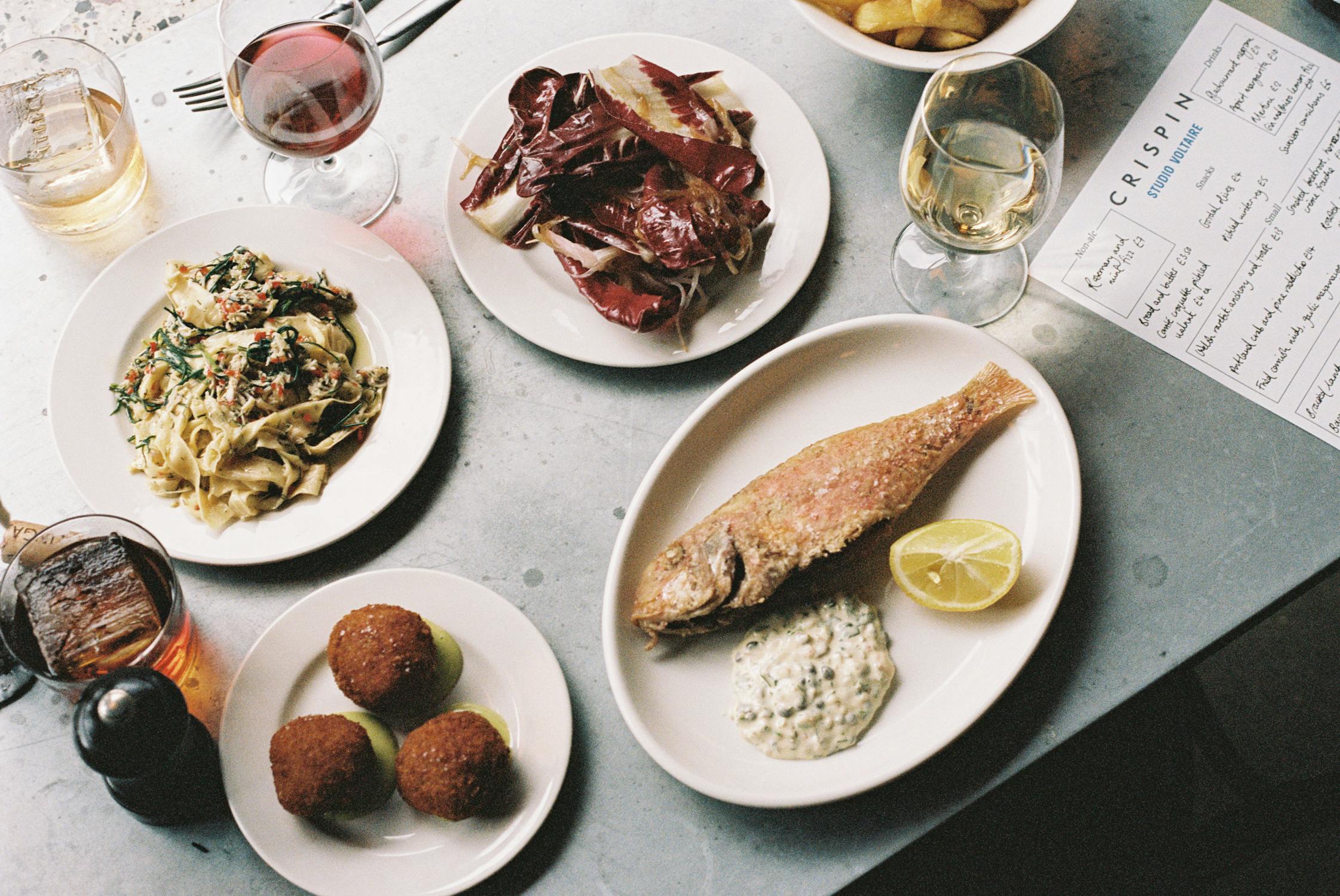 A photograph of a table with plates of food and drinks. There is a bowl of chips, a fried fish, croquettes, and different drinks that include red wine, a cocktail and a menu.