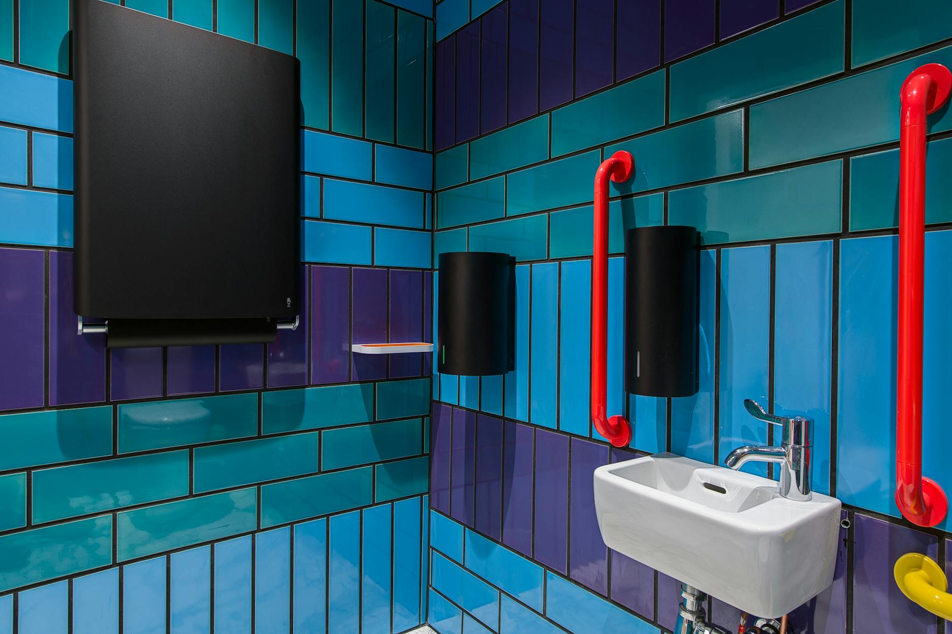 A colourful, tiled bathroom displays vibrant geometric patterns featuring circles, rectangles, and vivid hues of blue, pink, green, purple, and yellow. Two circular mirrors with sunburst designs are positioned on the wall, enhancing the whimsical aesthetic.