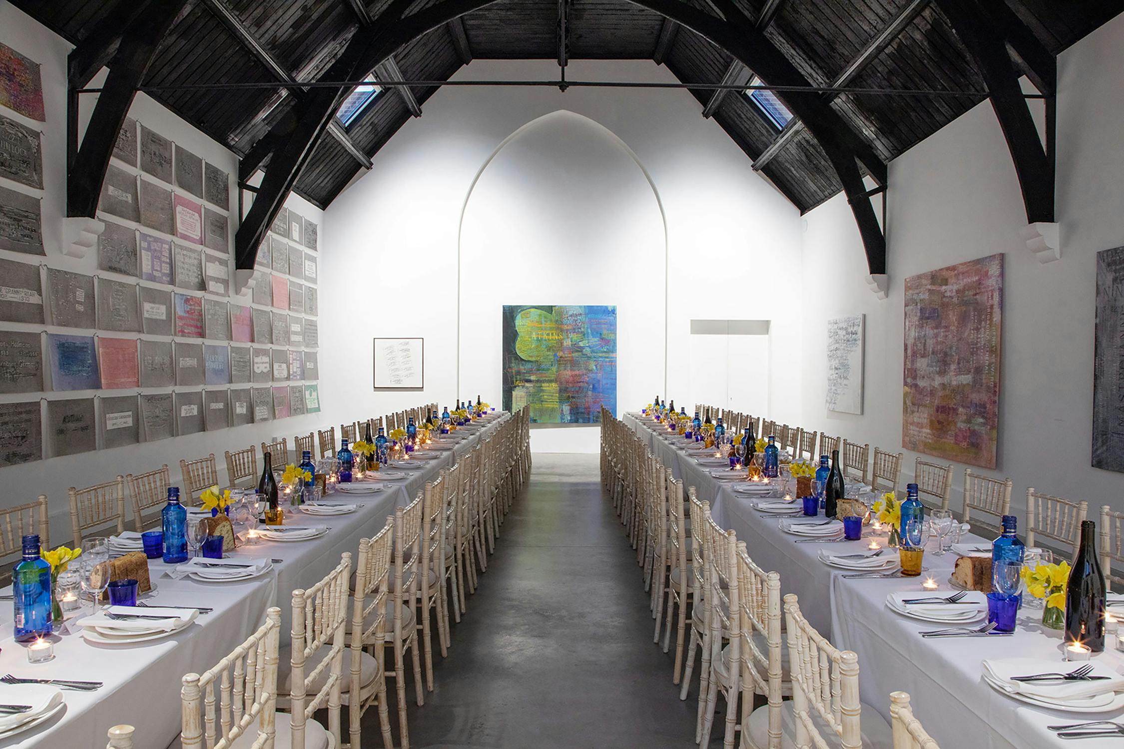 Studio Voltaire's vaulted gallery space featuring two long tables that span away from the camera. the tables are set with white table clothes, small bouquets of flowers, candles and colourful drinking glasses.