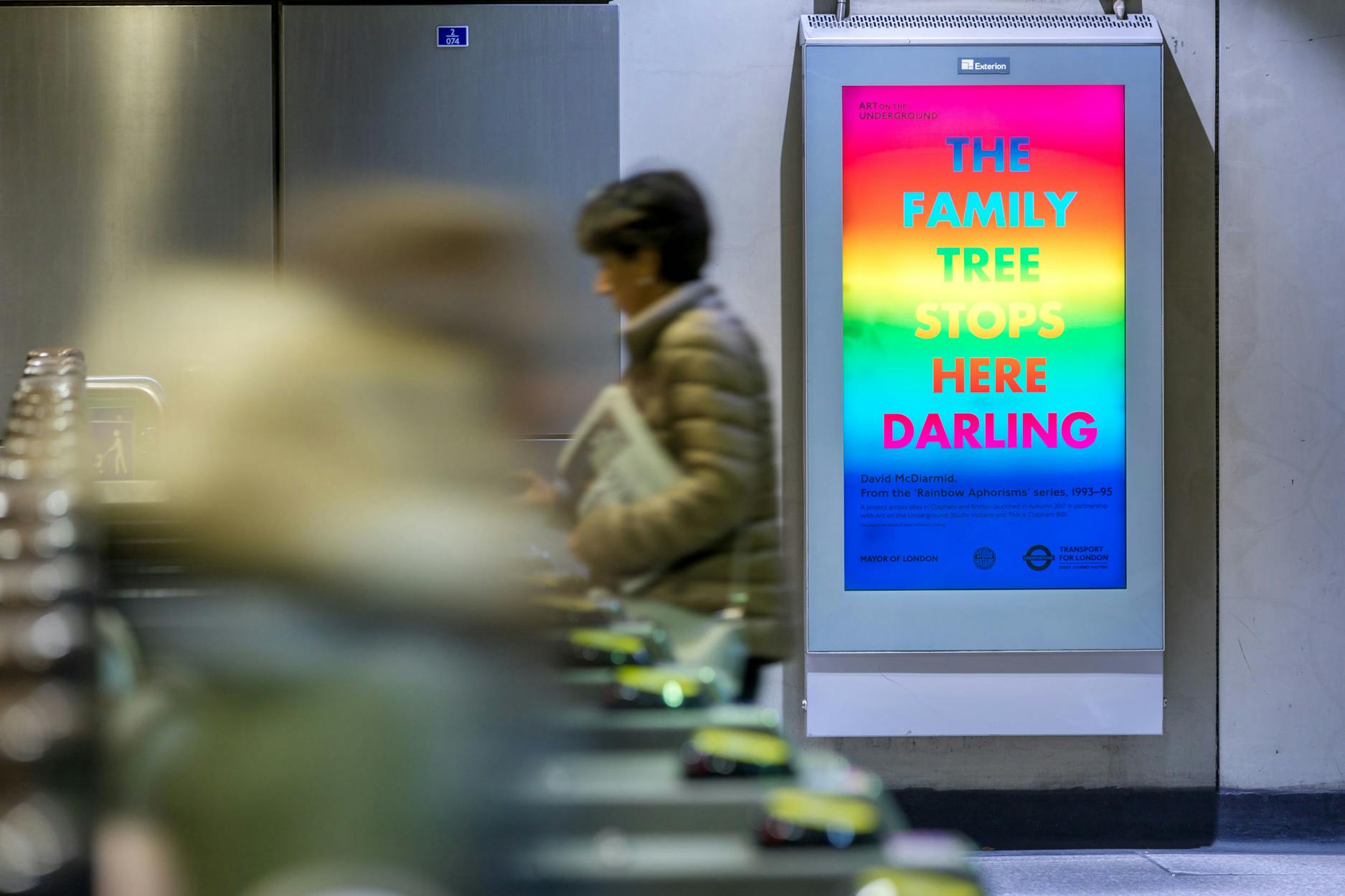 A colourful digital billboard reading “THE FAMILY TREE STOPS HERE DARLING” is displayed in a London Underground station as blurred commuters walk past turnstiles in the foreground.