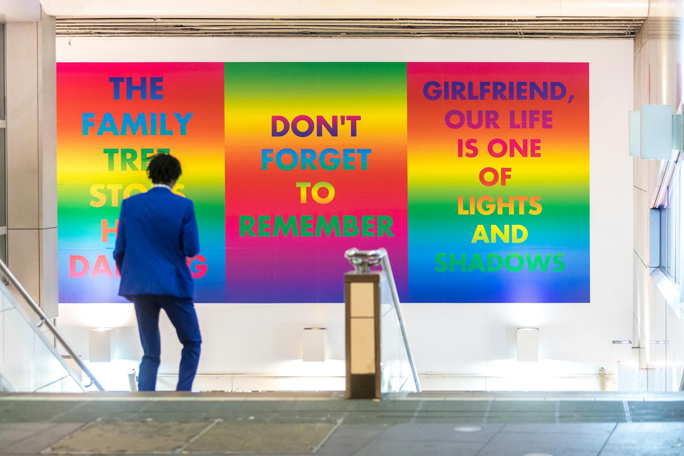 A person in a blue suit standing at the top of the stairs at the entrance of Brixton Underground station. A billboard above contains three rainbow panels displaying the phrases: "THE FAMILY TREE STOLE HIS DANCING," "DON'T FORGET TO REMEMBER," and "GIRLFRIEND, OUR LIFE IS ONE OF LIGHTS AND SHADOWS.