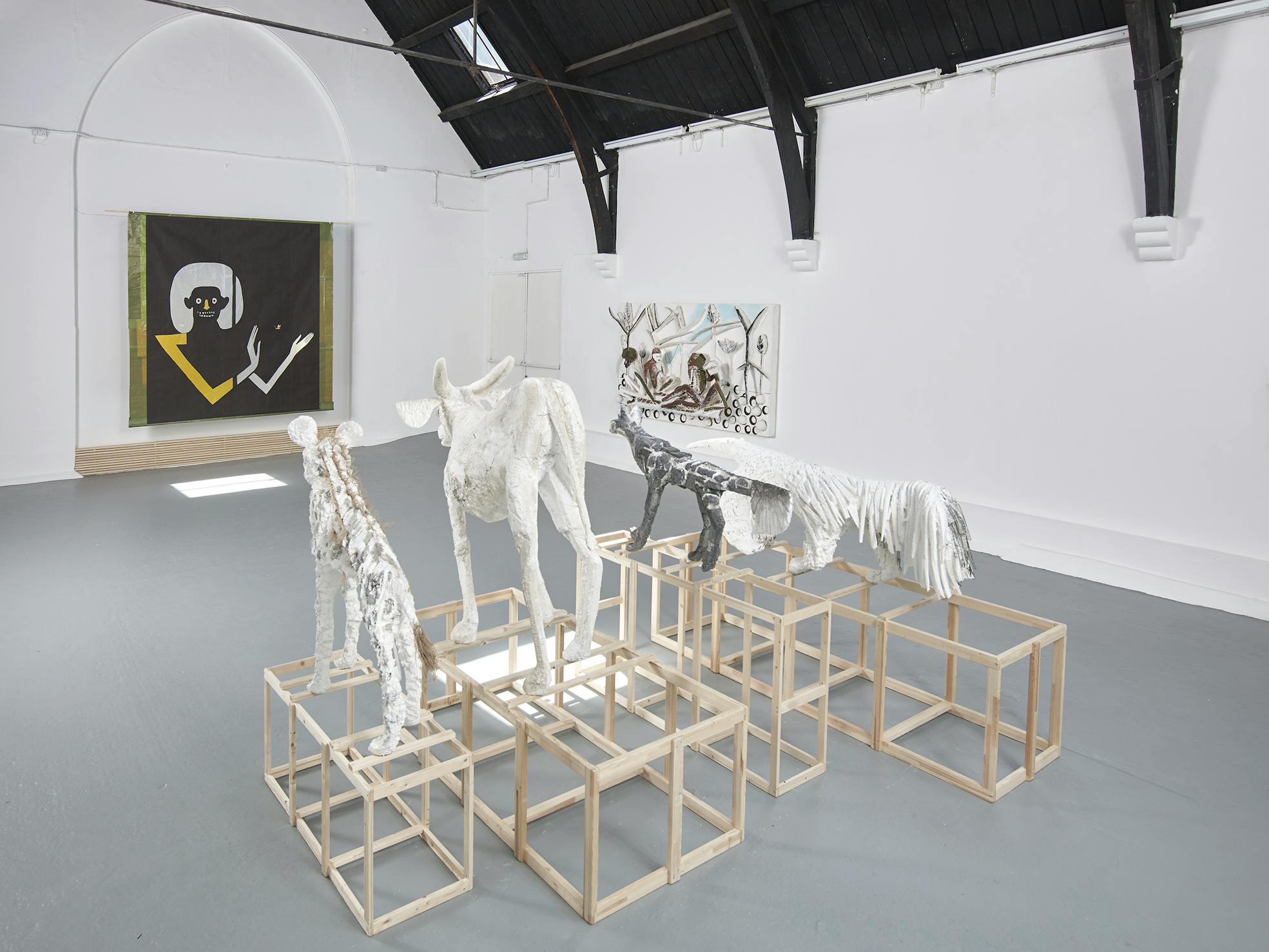 A white gallery features abstract animal sculptures on wooden cubes. The sculptures are covered in textured white and grey materials. On the wall, a large painting depicts a stylised figure in black, white, and yellow; another artwork hangs in the background.