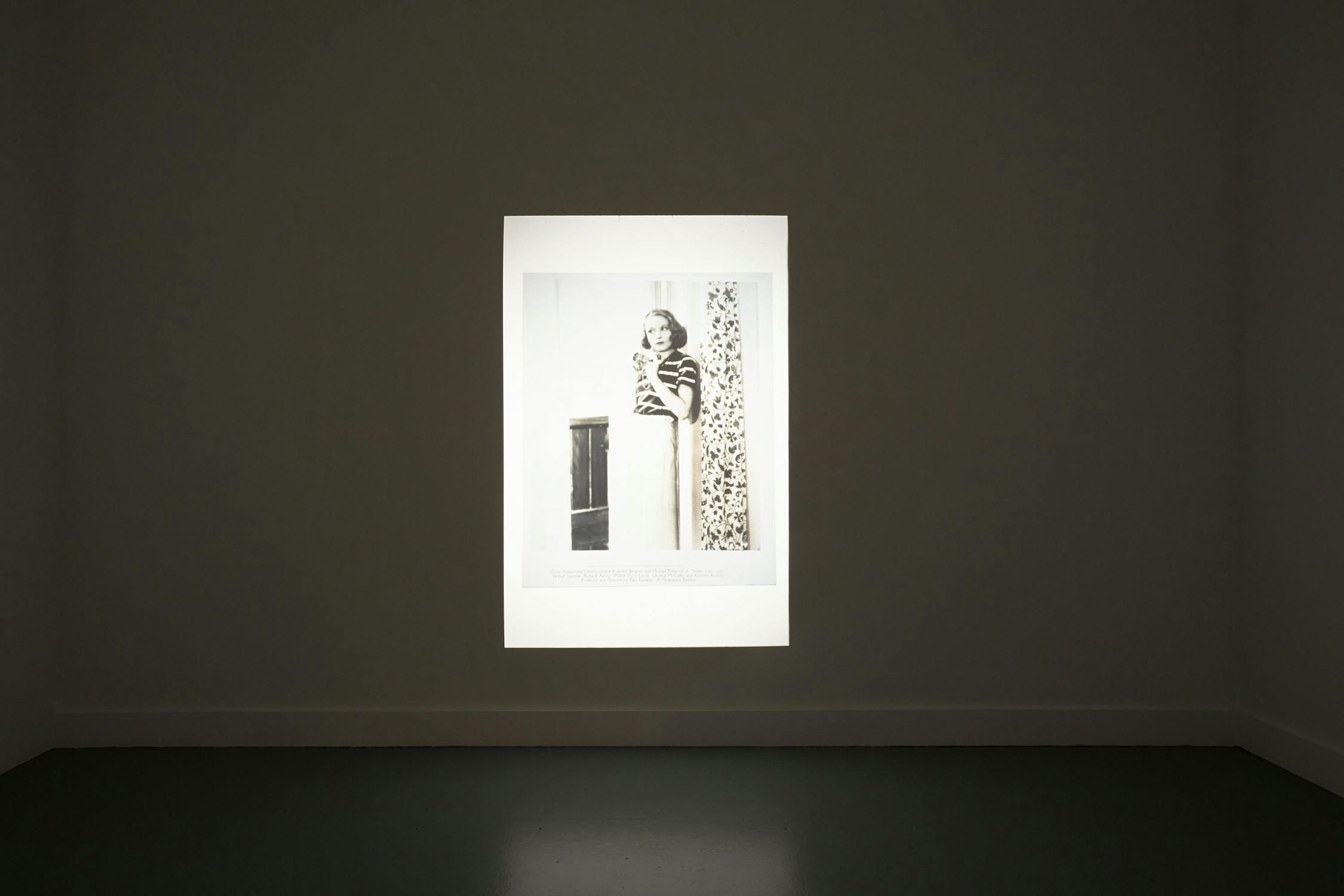 A photograph of a woman is illuminated and displayed on a gallery wall. The woman is standing beside a tall object with a patterned backdrop. The room is minimally decorated with the focus on the central image. The lighting emphasises the displayed photograph.