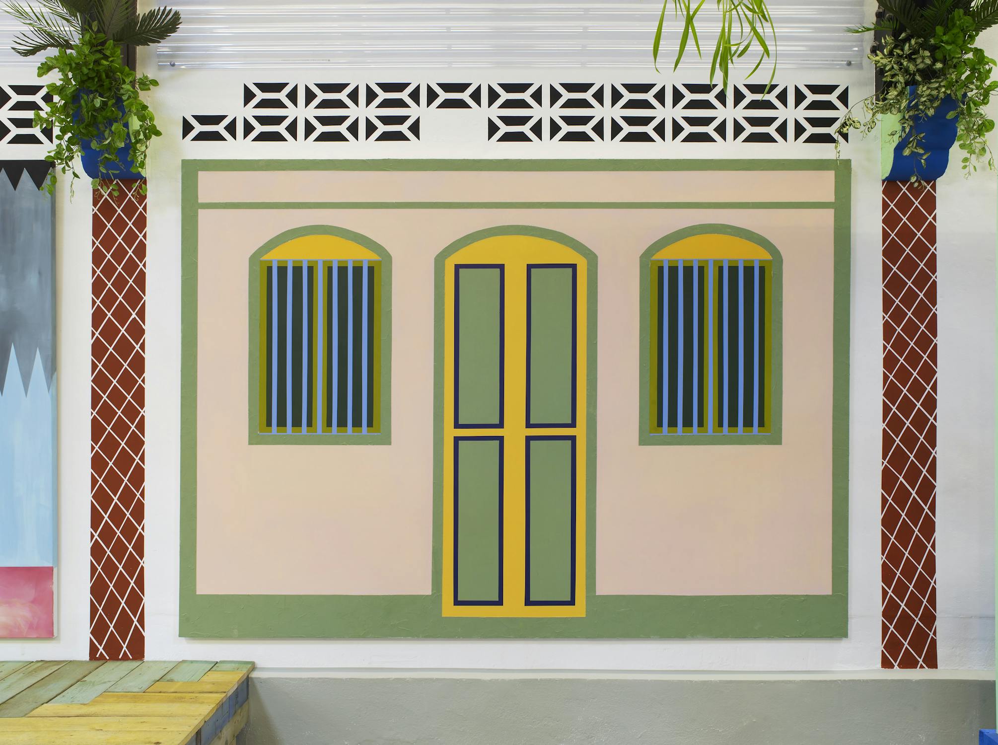 A detail from Sol Calero’s installation at Studio Voltaire, featuring a wall designed to resemble a stylised house façade. It includes two arched, blue-barred windows and a central door, all outlined in green and yellow, with geometric vents and hanging potted plants completing the decorative scene.