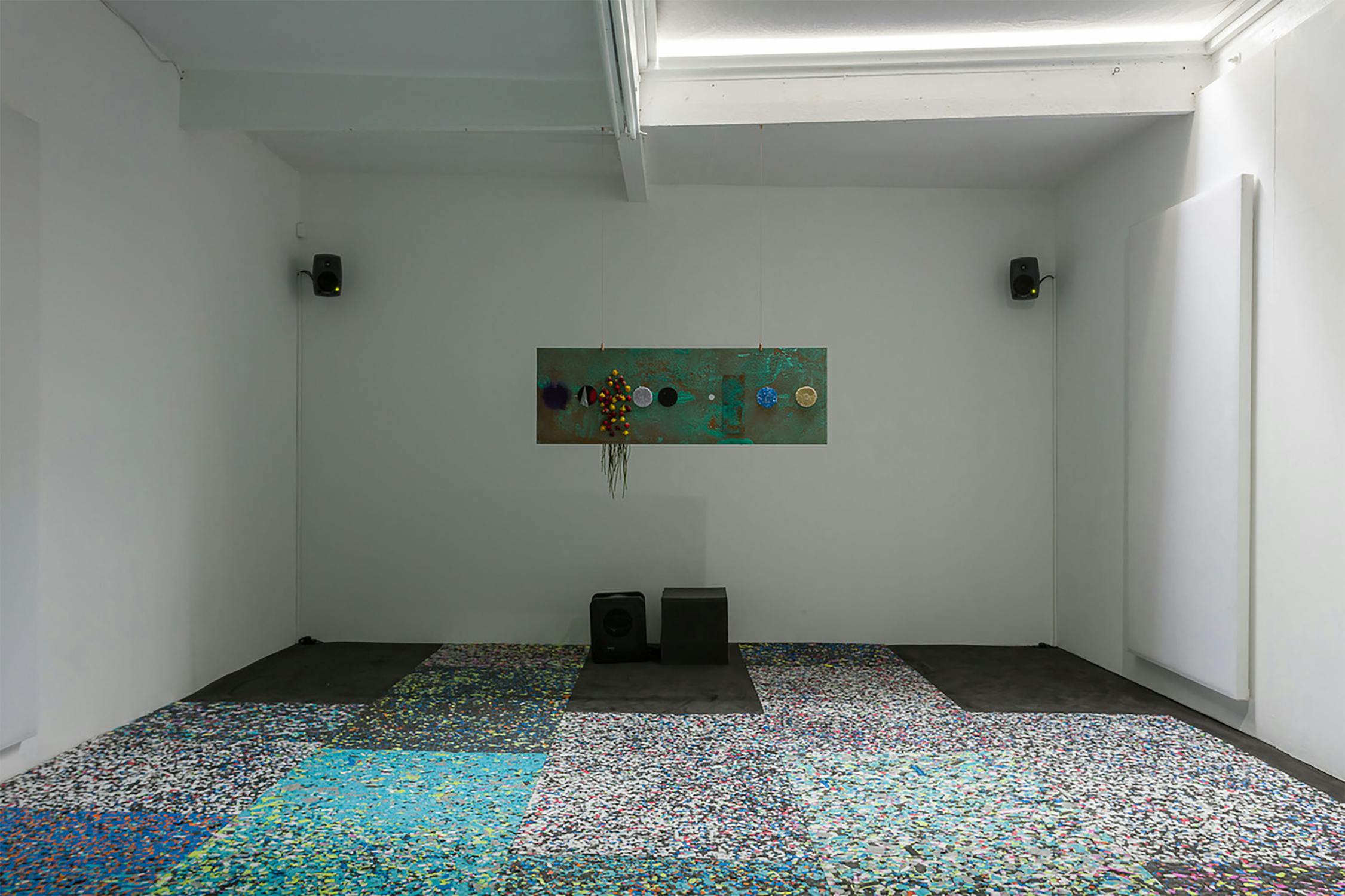 A minimalist gallery room with white walls features a colorful abstract artwork centered on the back wall. The floor is covered with a vibrant, multicolored mosaic carpet. Speakers are mounted on the walls, and two rectangular objects sit on the floor.