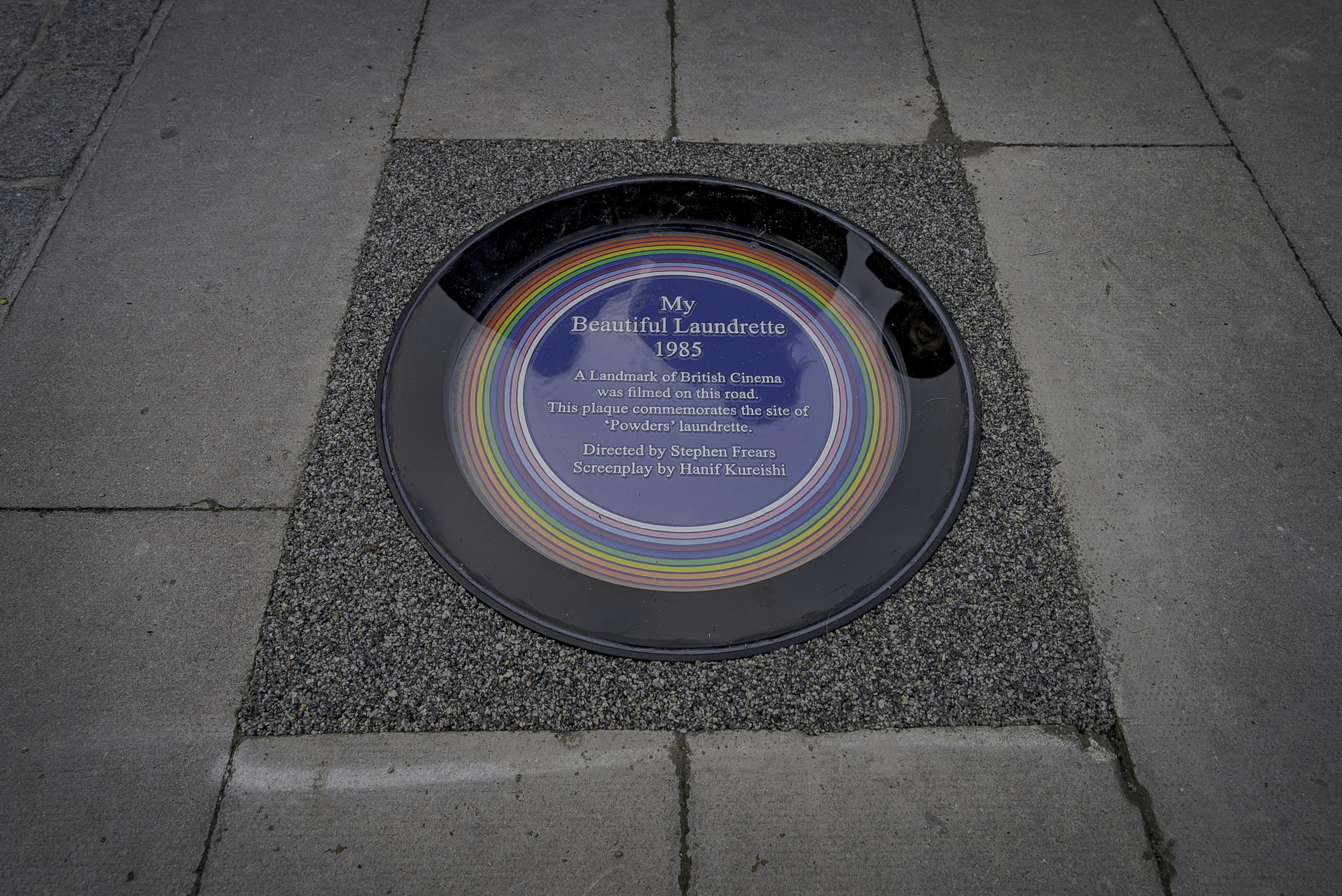 A circular plaque set into a pavement, reading: “My Beautiful Laundrette 1985. A Landmark of British Cinema was filmed on this road. This plaque commemorates the site of Powder’s laundrette. Directed by Stephen Frears. Screenplay by Hanif Kureishi.”.