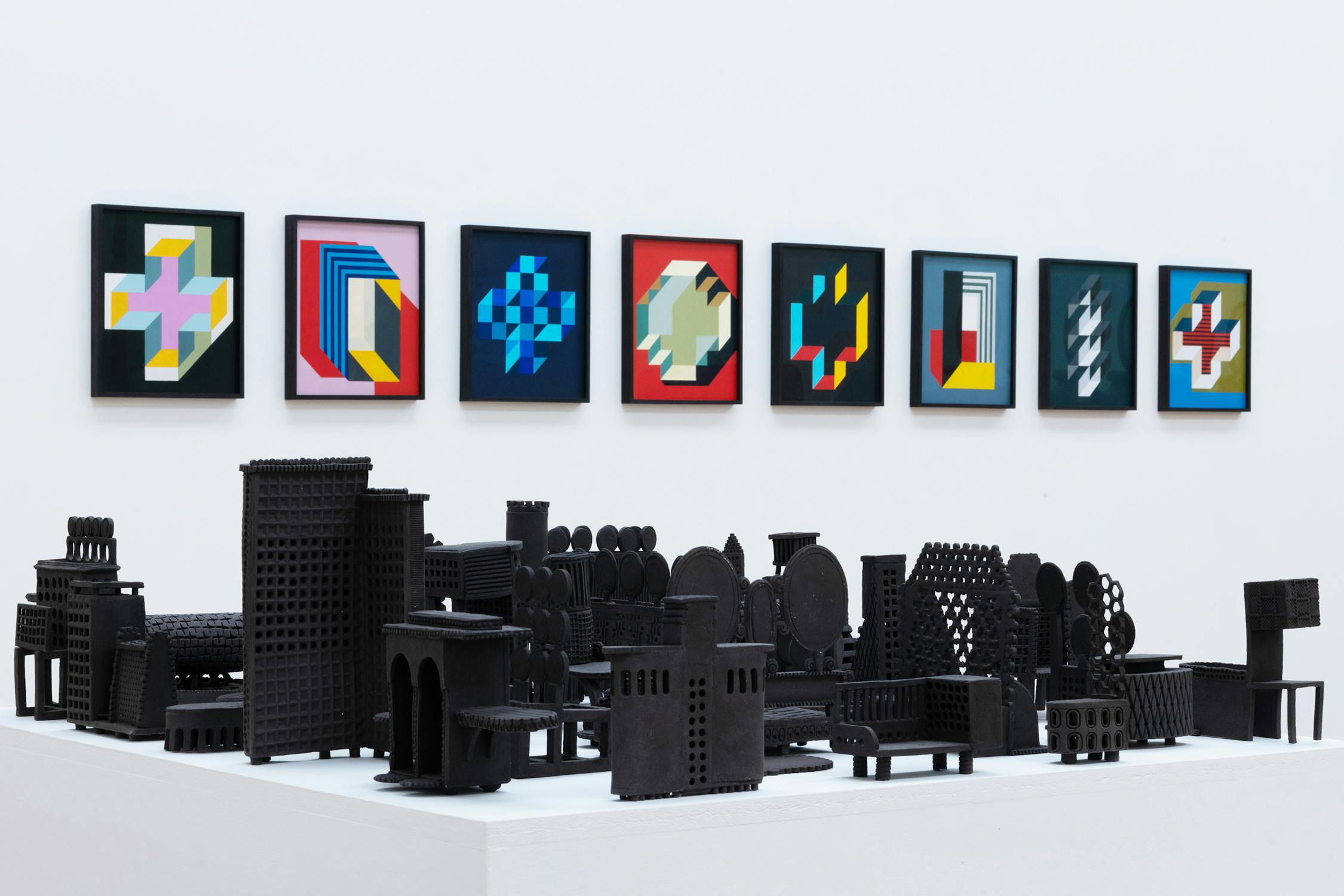 A series of abstract geometric paintings in colorful frames are displayed on a white wall. In the foreground, a collection of black sculptural models resembling stylized buildings is arranged on a white platform.