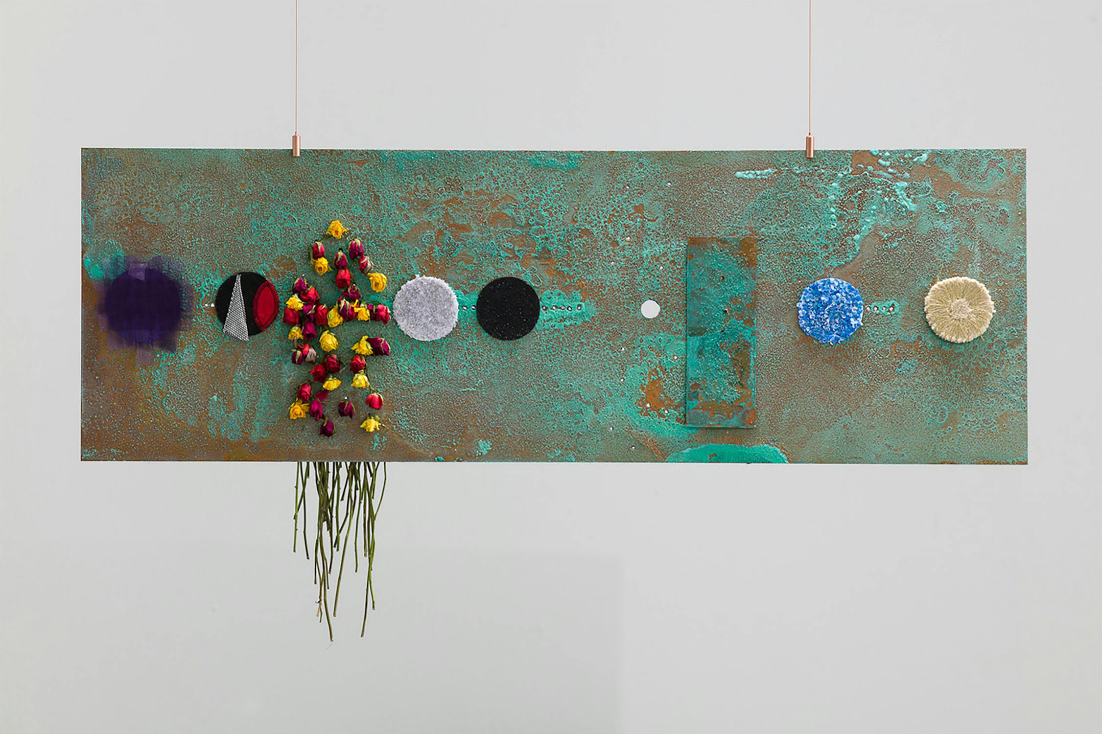 A rectangular artwork with a textured, greenish surface features various circular elements, including colorful flowers, patterns, and woven designs. Thin strips hang from two points, and foliage dangles from one section.