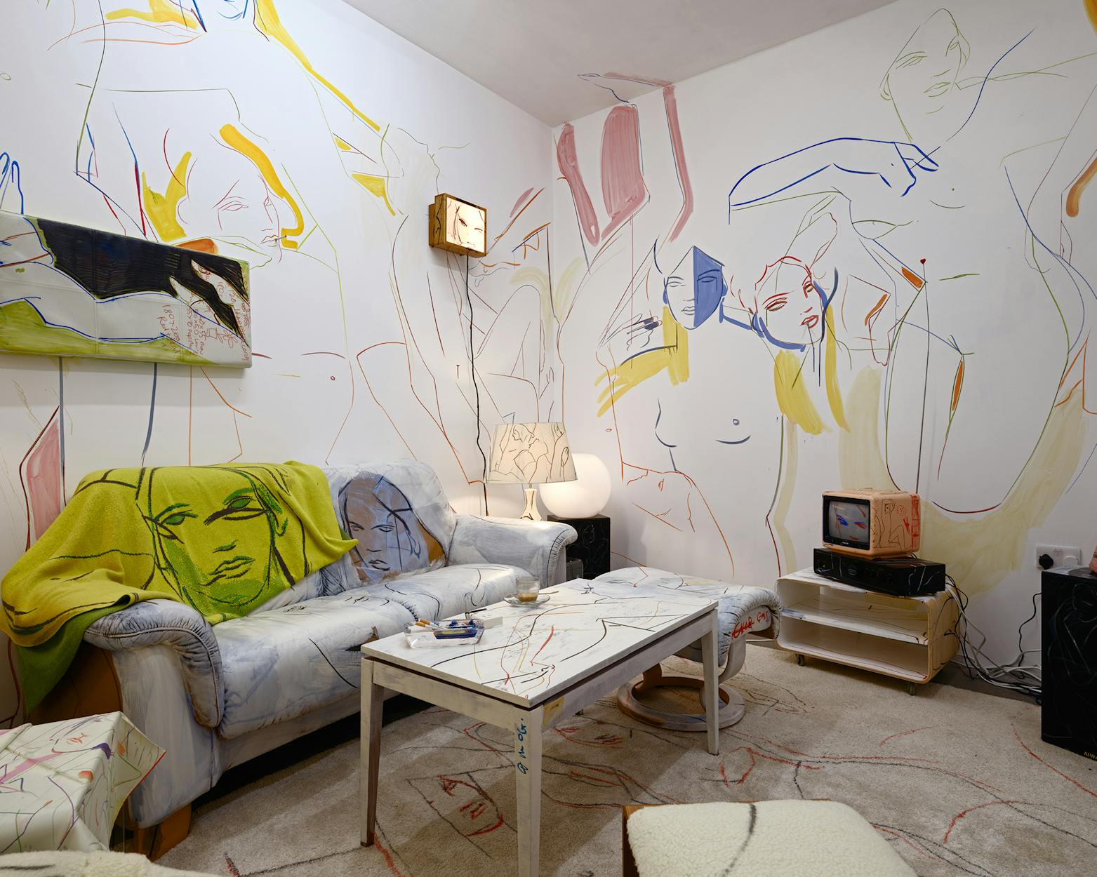 A living room installation by France-Lise McGurn with abstract line drawings of colourful human figures covering the walls, furniture, and décor. The space features a couch, coffee table, lamp, small TV, and unique wall art, all incorporating the same artistic style.