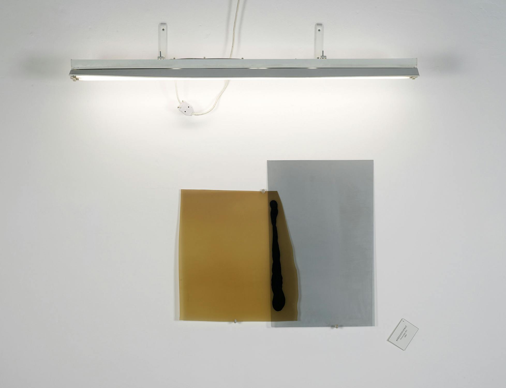 A fluorescent light fixture illuminates two abstract translucent sheets, one yellow and one grey, displayed on a white wall.