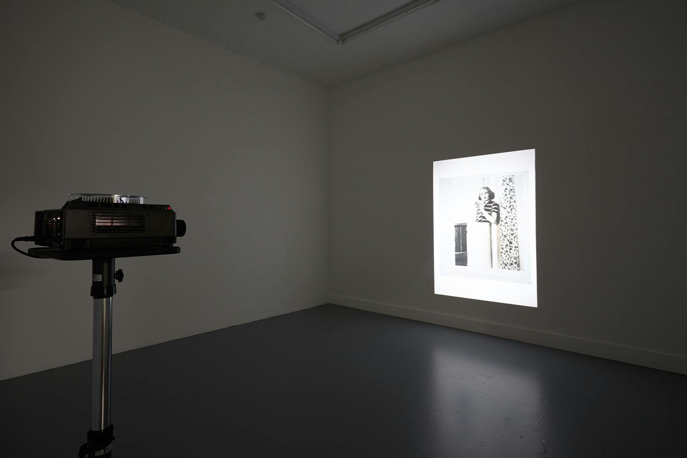 A dimly lit room with a slide projector on a stand, projecting a black-and-white image onto a white wall. The projected image appears to be of a piece of art or a photograph, featuring abstract shapes and patterns. The floor and walls are plain and grey.