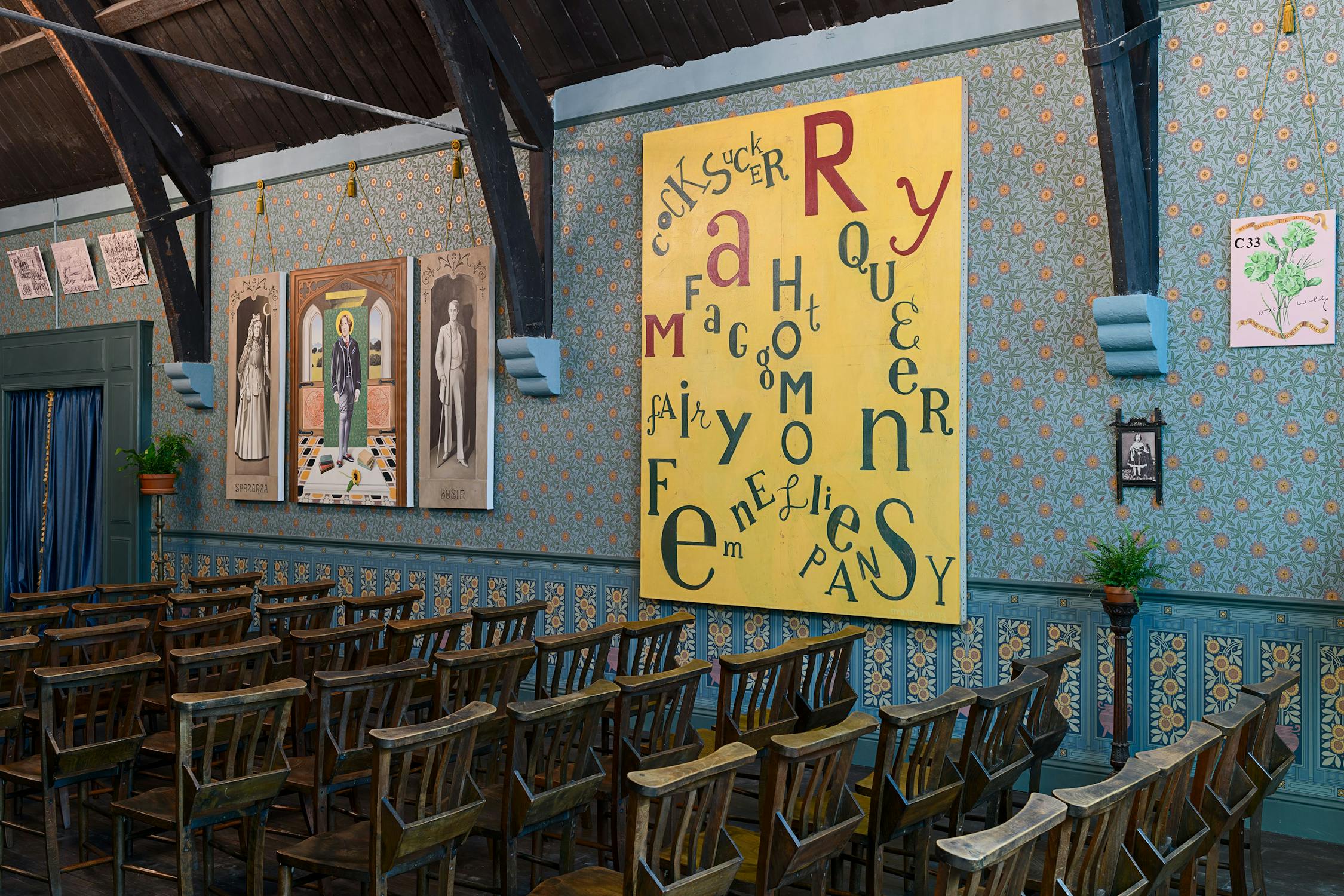 Rows of empty wooden chairs face large colourful artworks on patterned blue wallpaper. A yellow poster with words like "Queer," "Fem," "Homosexual" in playful fonts is prominent. Potted plants and framed pictures decorate the room. The ceiling is dark wood.