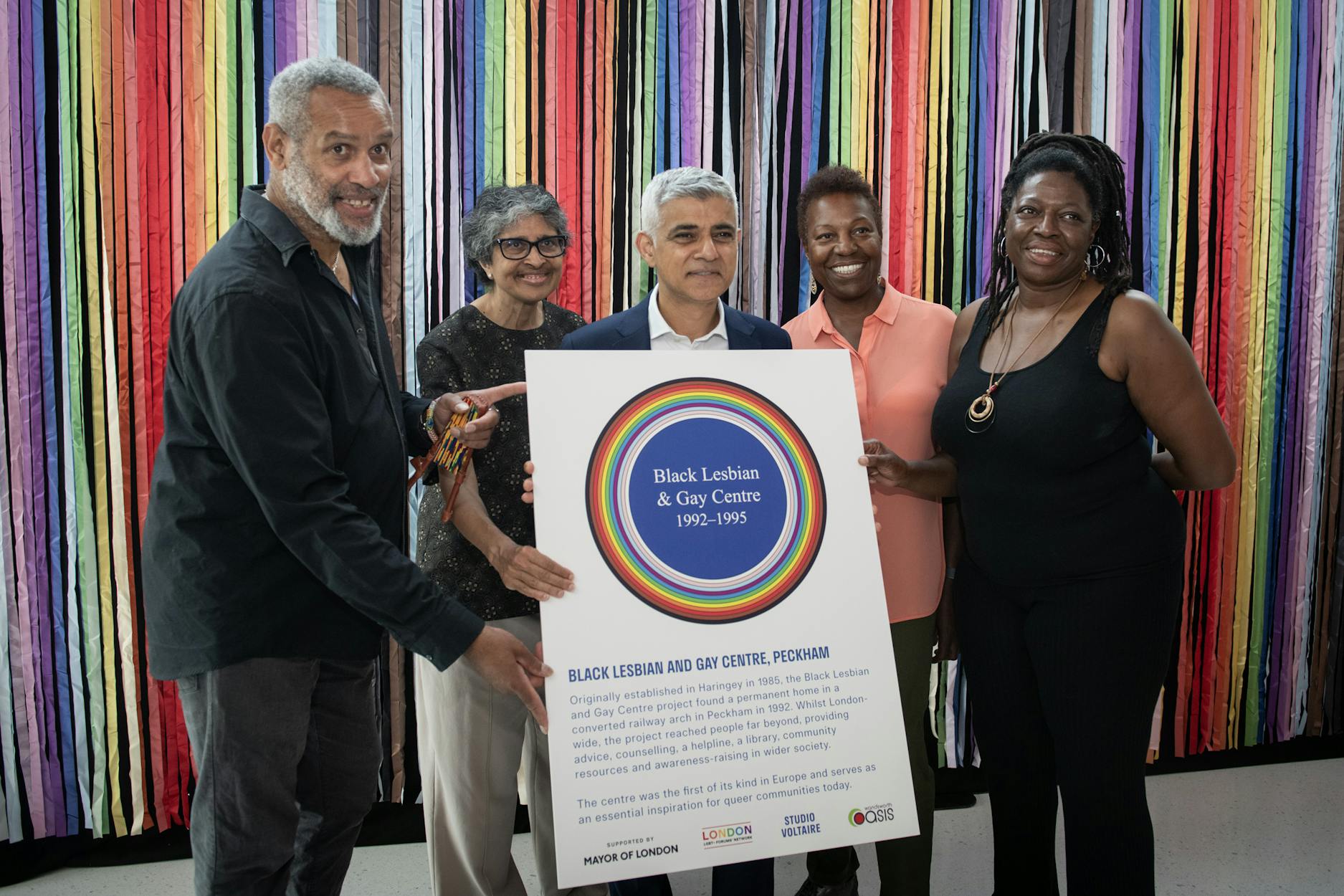 Five people stand smiling in front of colourful hanging ribbons, holding a sign that reads “Black Lesbian & Gay Centre 1992–1995” and provides information about the centre’s history in Peckham, London.