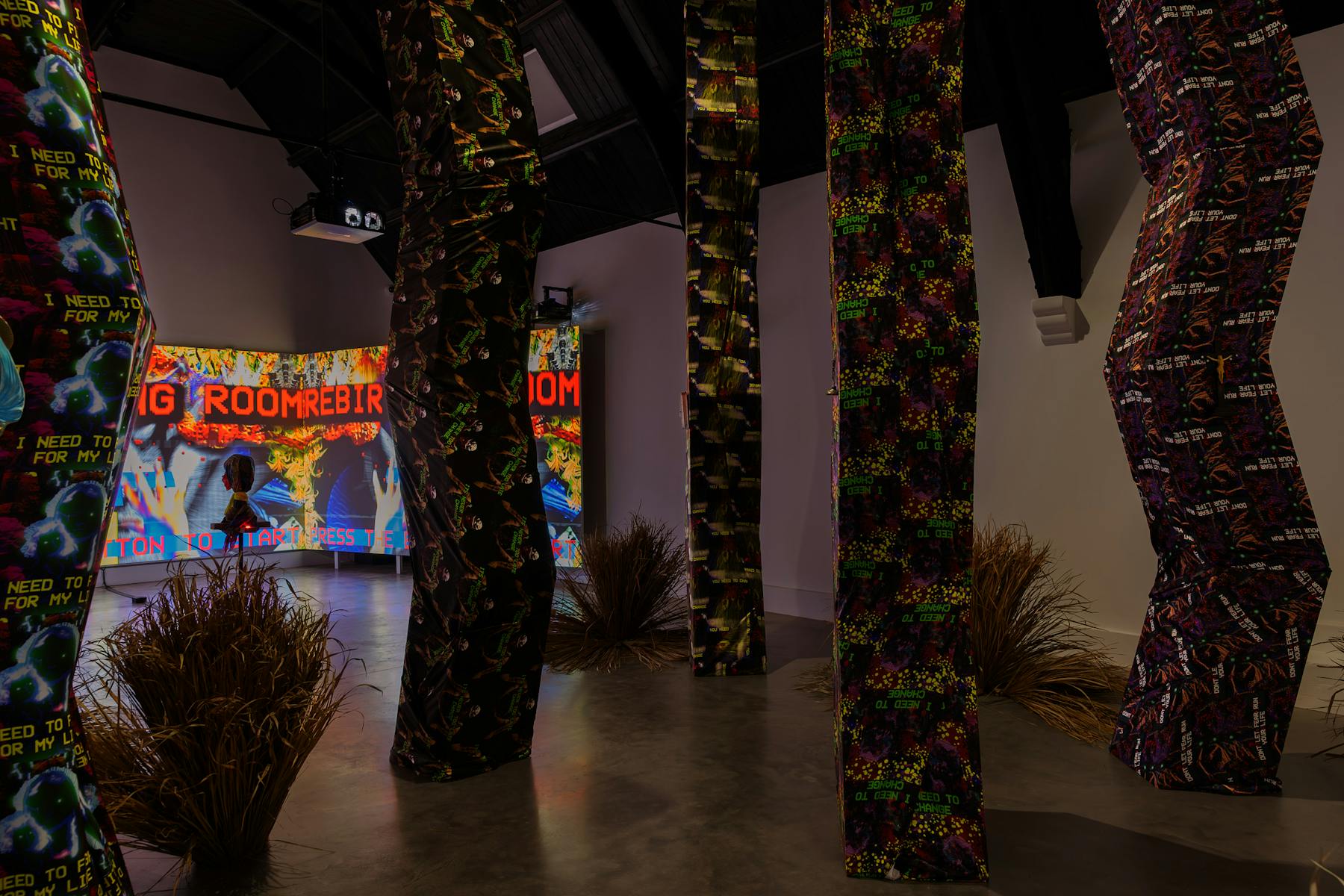 A photograph of a dimly lit room, that is filled with tall angular structures that reach from the floor to the ceiling. The structures are wrapped in vibrantly patterned fabrics. In the bcakground, through the structure, viewers can see a large screen with colourful projections.
