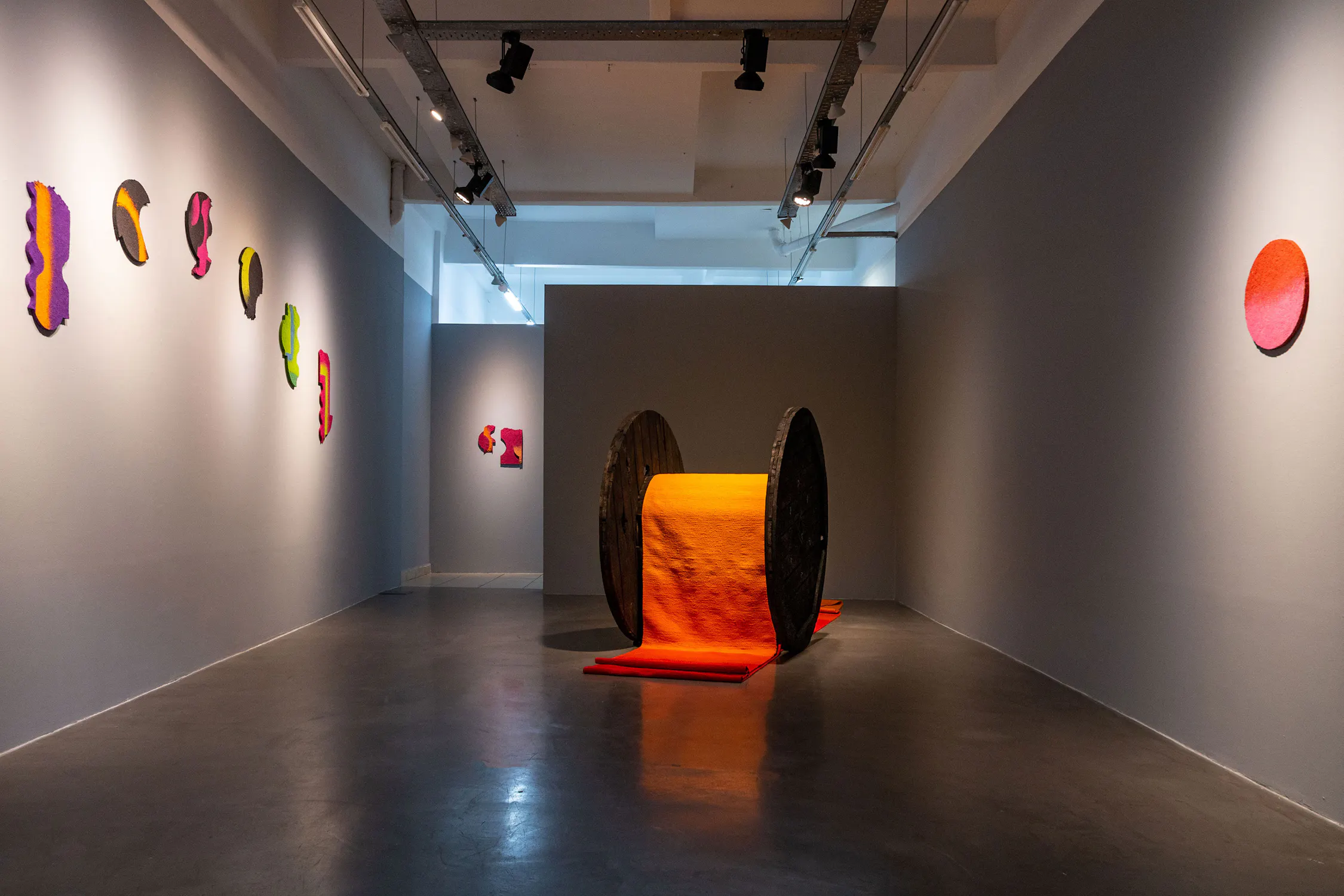 A gallery with grey walls displays colourful, abstract art. At the centre, a giant spool with bright orange thread unrolls onto the floor. Abstract, vibrant pieces hang on both side walls and the back wall. Track lights illuminate the space.