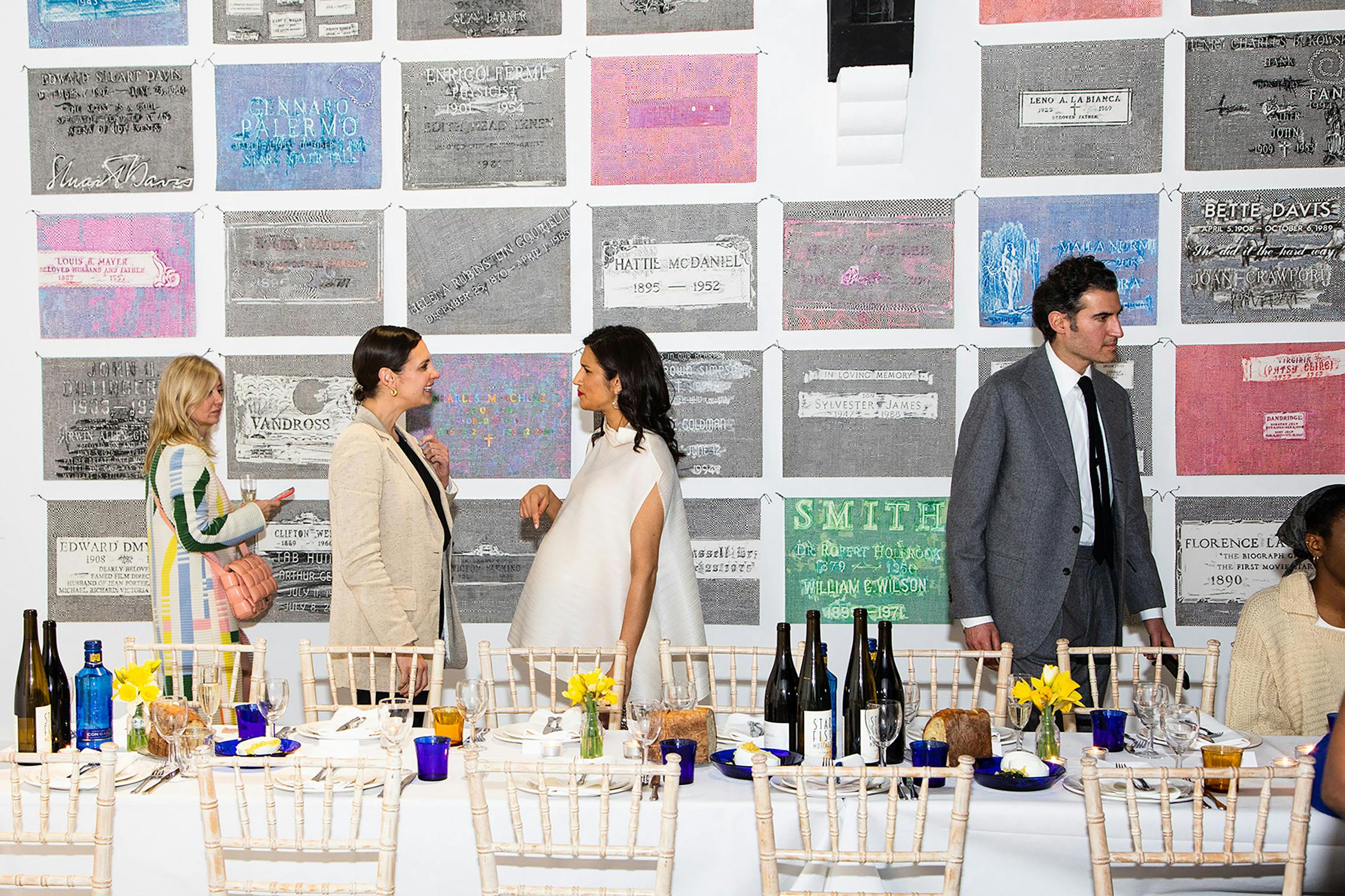 Dinner party guests speaking to one another at a long table with a white table cloth, bottles of wine, small bouquets of flowers. the quests stand in front of a wall adorned with colourful square artworks.