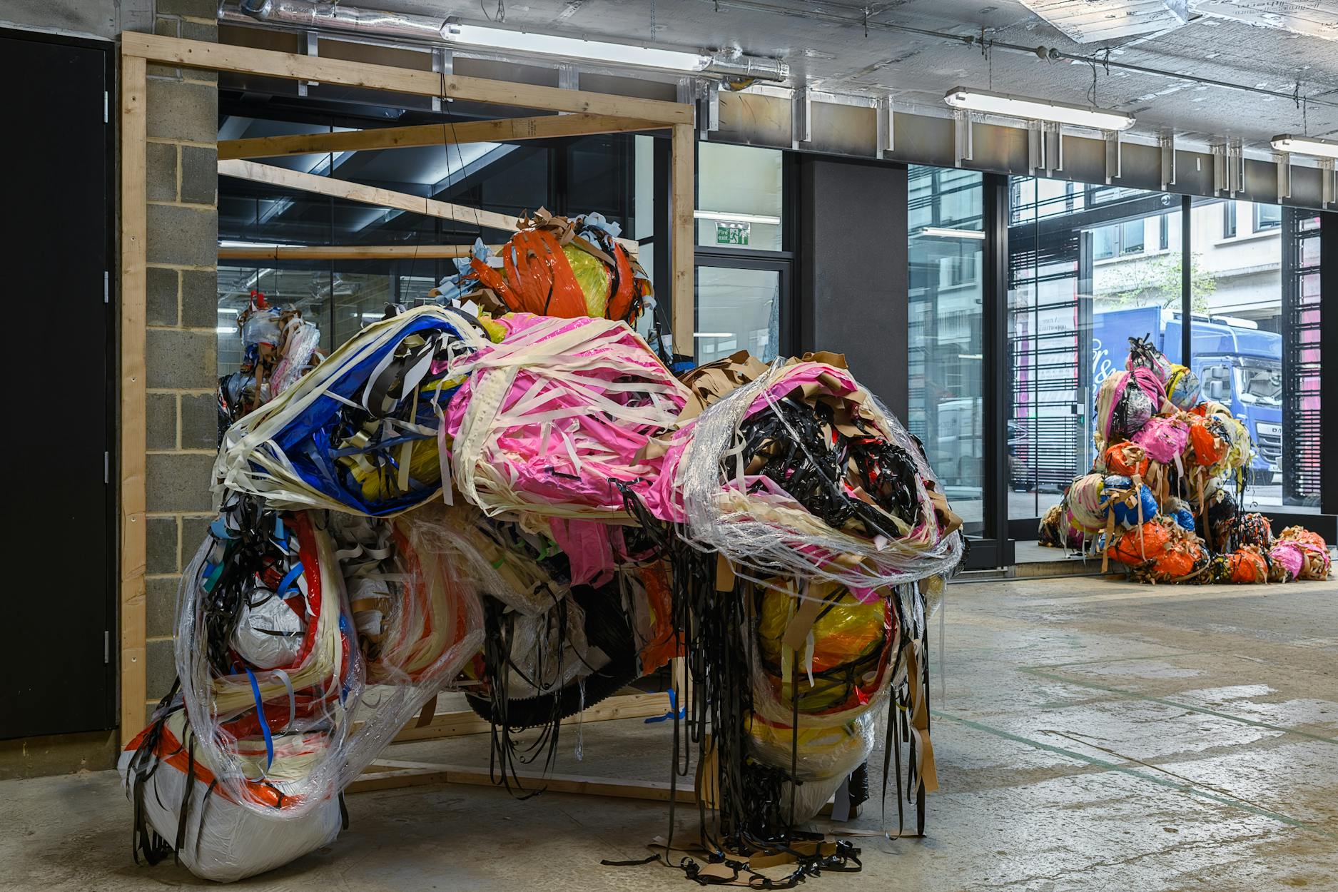 A large indoor sculpture by Nnena Kalu, made from colourful, tangled plastic strips, ropes, and assorted materials forms an abstract shape on a concrete floor. In the background, a similar, smaller sculpture is visible near large windows letting in natural light.