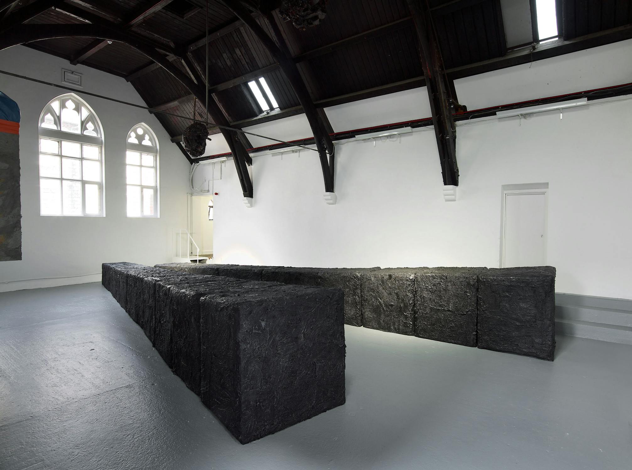 A gallery with high, dark wood ceilings and arched windows features a row of large, rough-textured black rectangular blocks arranged in a zigzag pattern on a grey floor, contrasting with the bright white walls and natural light.
