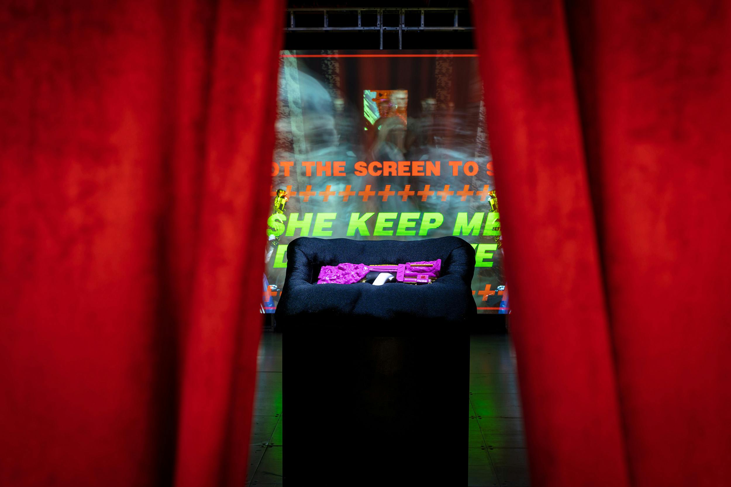 A pink machine gun on top of a black pillow upon a black plinth seen through red curtains