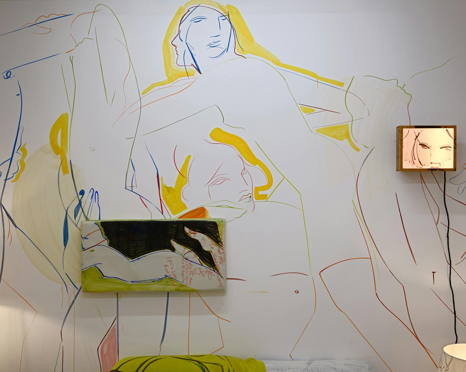 A wall features large, abstract painted mural of nude human figures in pastel colours, with two framed artworks—one rectangular and one square—hung on top, partially blending into the mural.