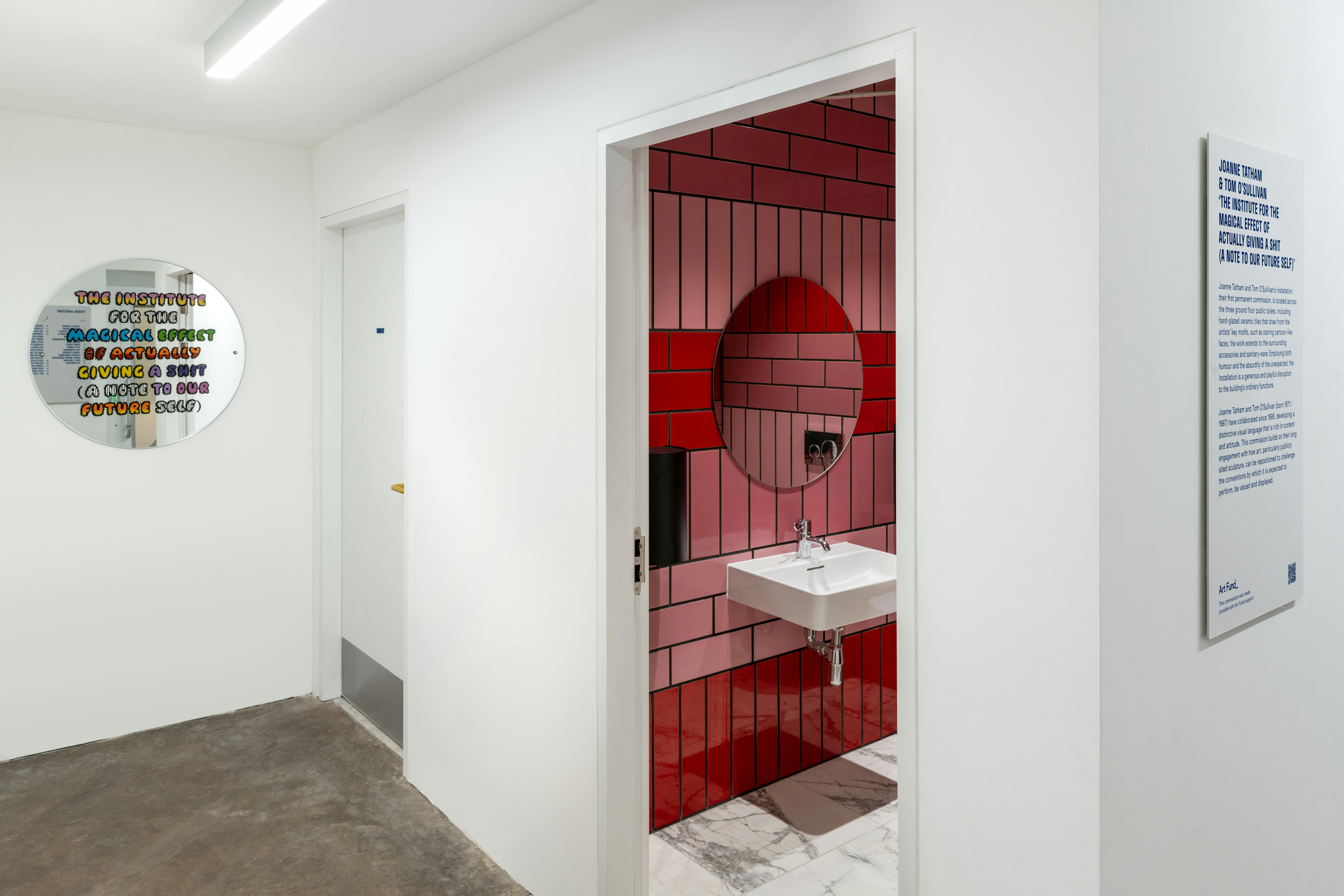 A white hallway with a restroom door open, revealing a bathroom decorated with red and pink tile walls and a white sink. A round mirror on the left wall displays colourful text. Nearby, a plaque with writing is mounted on a white wall.