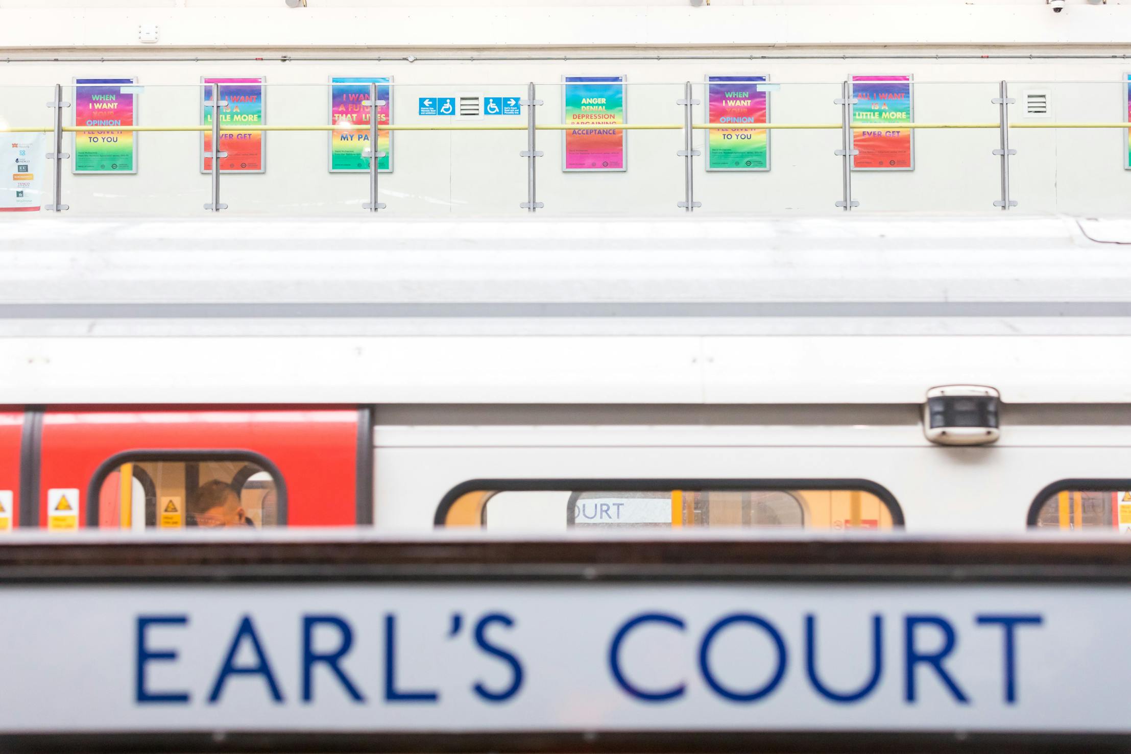 A London Underground train at Earl’s Court station, with a platform sign in the foreground. Colourful rainbow-gradient posters by David McDiarmid can be seen on the wall above the train in the background.
