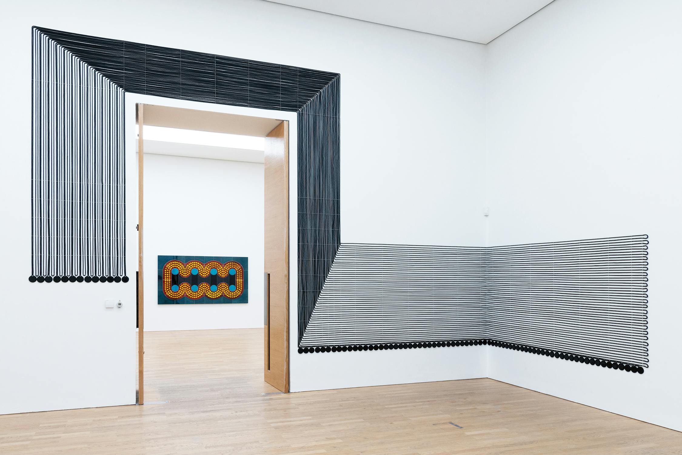 A modern art installation by Lubna Chowdhary with black lines creating geometric shapes on white walls. Some lines flow around an open doorway, leading to another room displaying a colorful abstract painting. The floor is light wood.