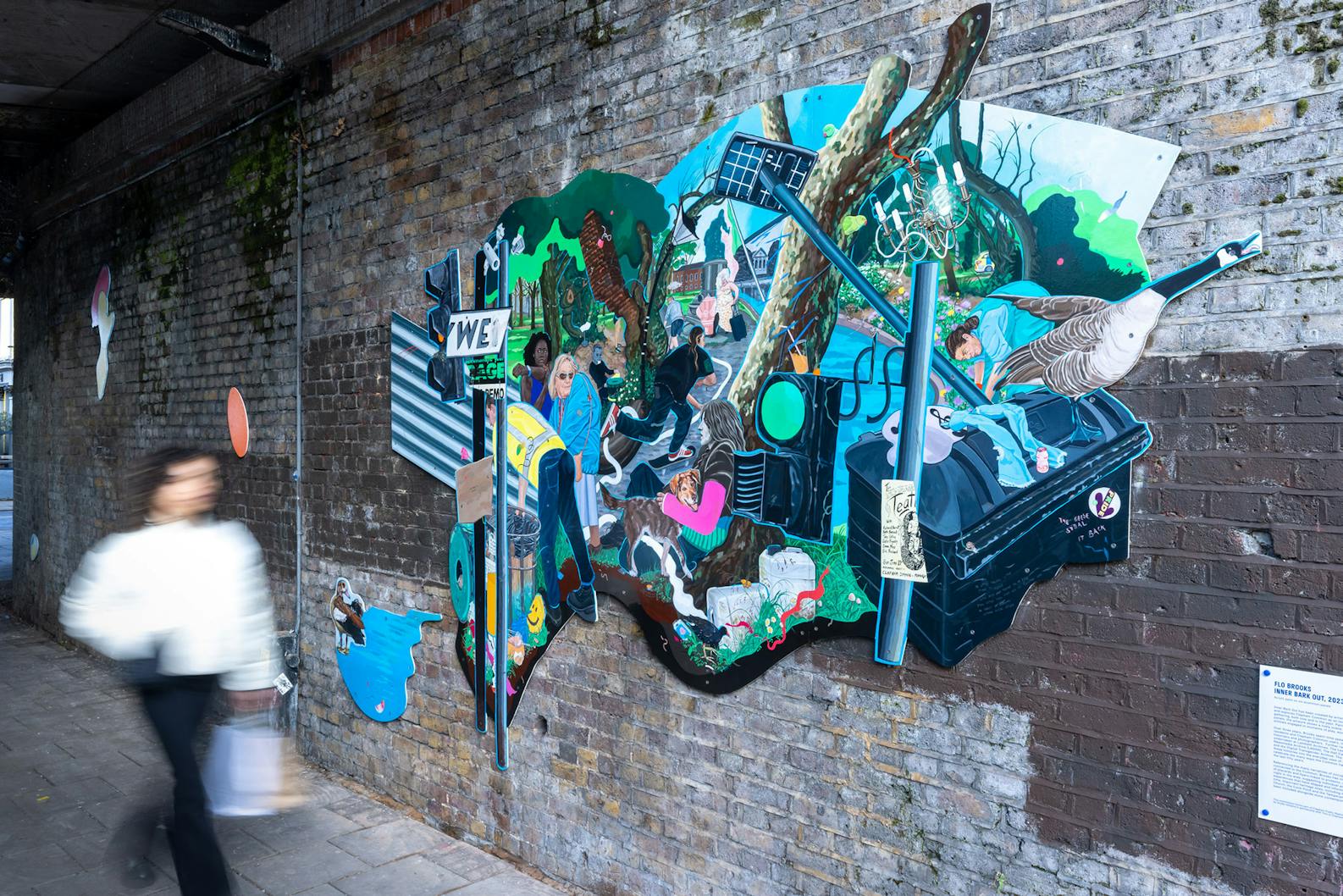 A vibrant and eclectic mural by Flo Brooks is painted on a brick wall. The mural features various elements like a man holding a baby, wildlife such as birds, and urban imagery including street signs and graffiti. There's also a small plaque with text on the wall beside the mural.