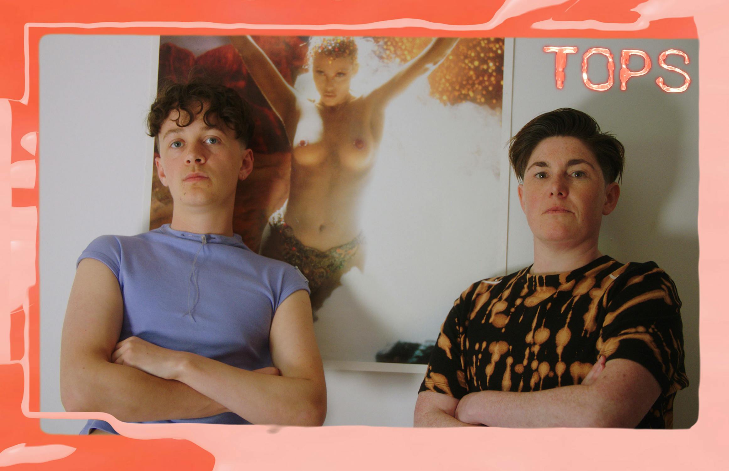 Two people stand with arms crossed in front of a framed photo of a woman. A decorative pink border surrounds the image with the word "TOPS" in the top right corner.