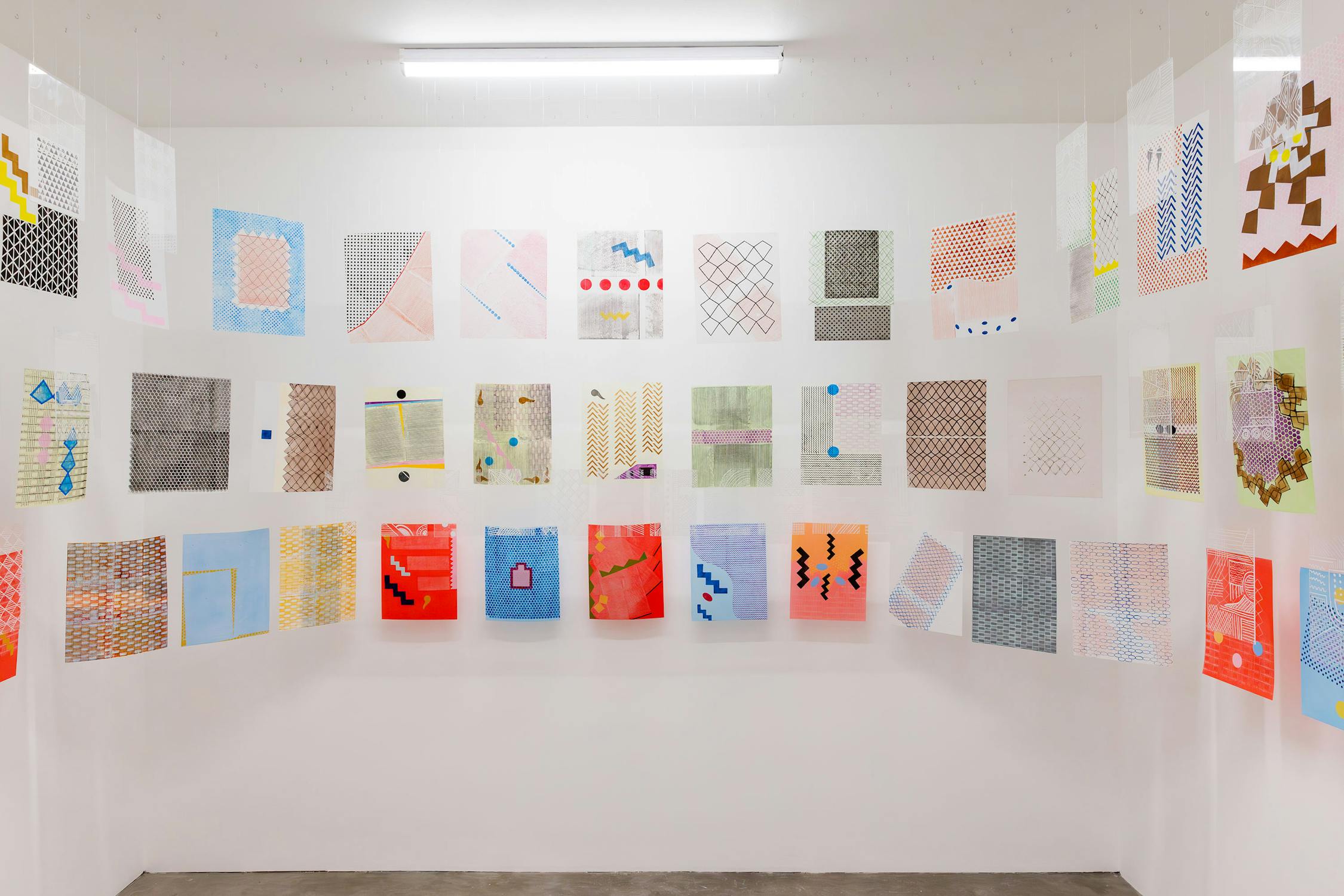 A white room with colourful abstract artworks by Bamidele Awoyemi and Adia Wahid displayed on three walls. The pieces feature geometric patterns, lines, and shapes in various hues, creating a vibrant and eclectic atmosphere. A fluorescent light illuminates the display.