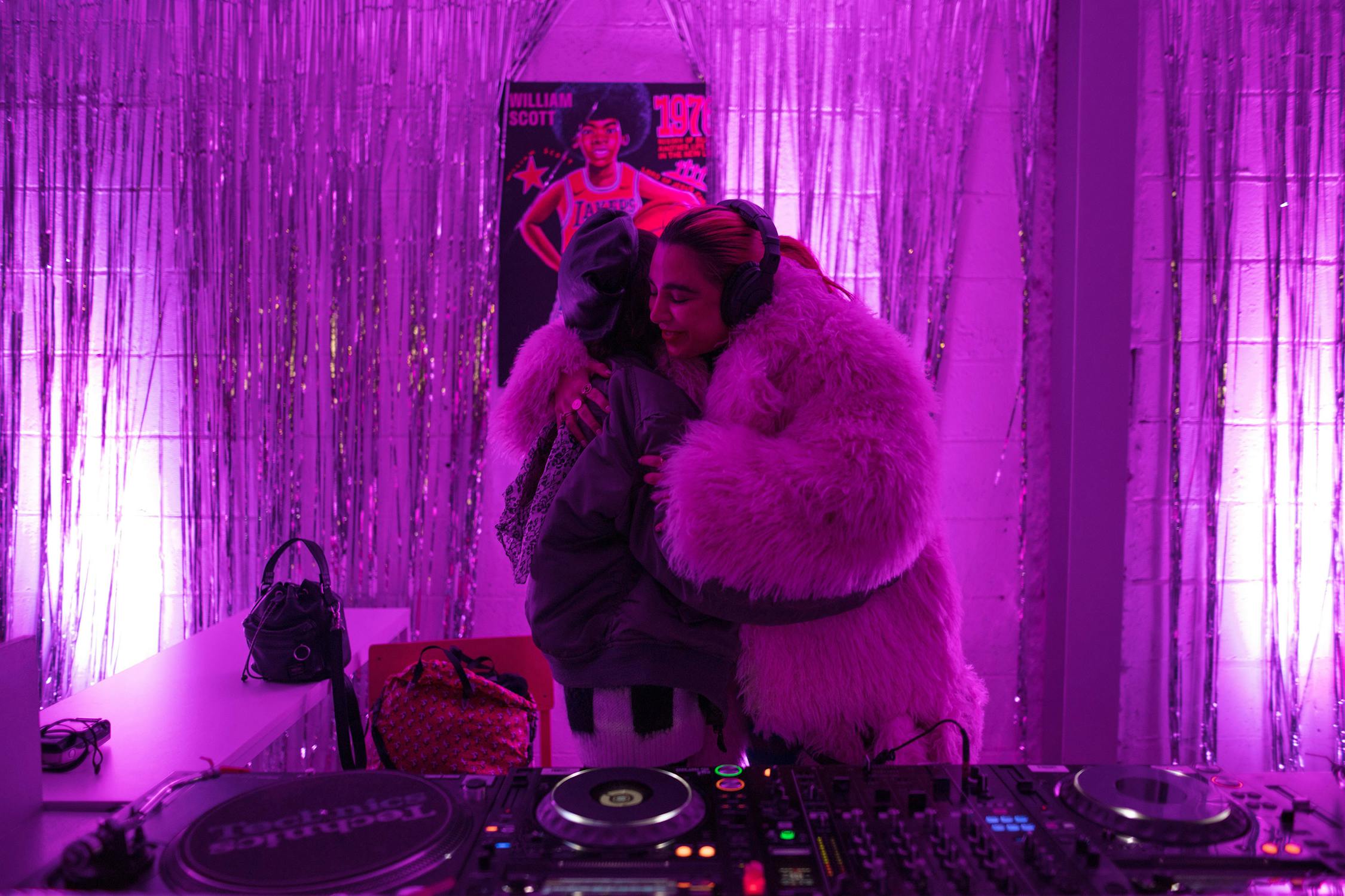 Two people hug in a room decorated with shiny silver streamers and purple lighting. One wears a large pink fur coat and earmuffs; both have bags near them. A Lakers poster by William Scott is on the wall in the background.