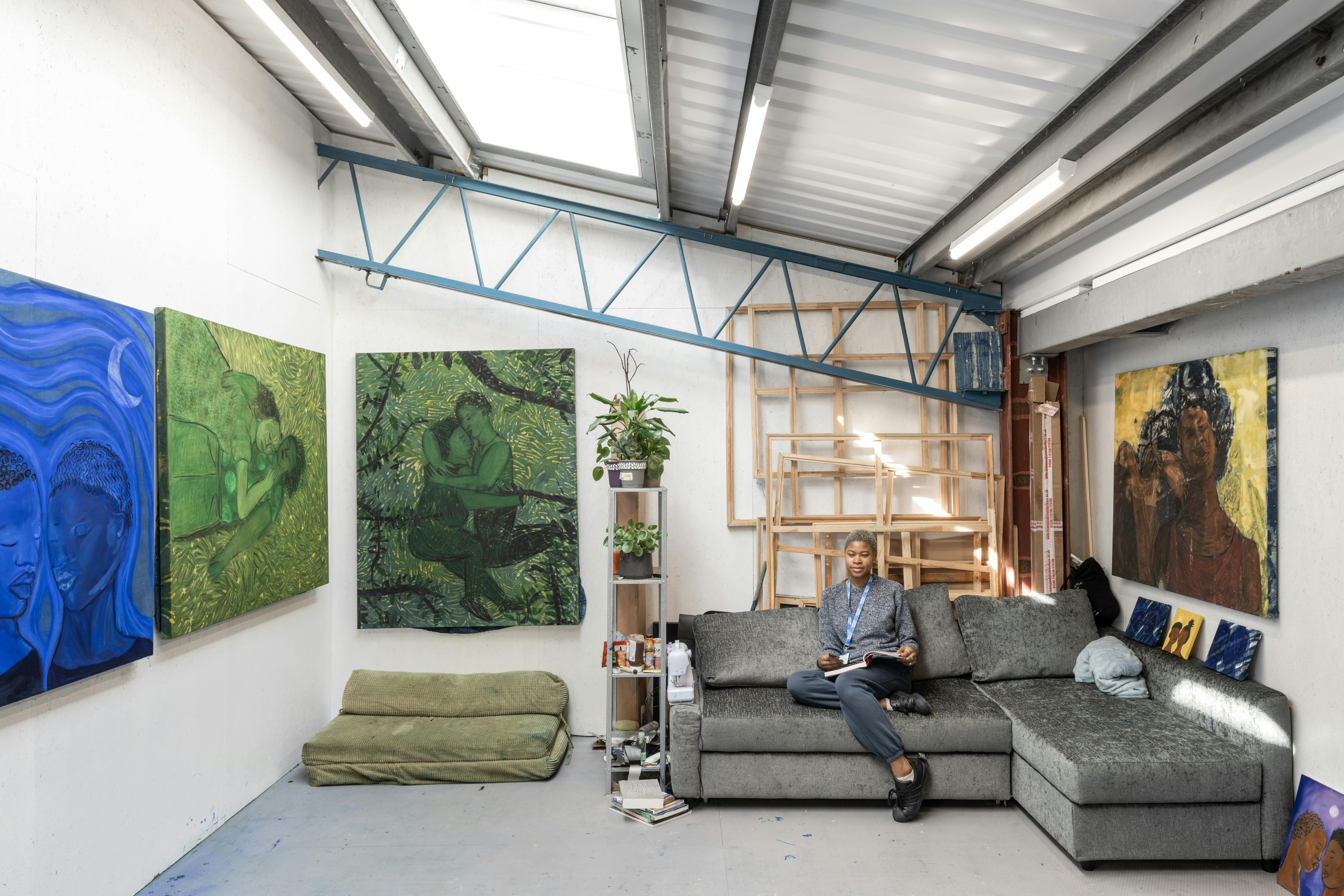 A person sits on a grey sectional sofa in an art studio with white walls, surrounded by large colourful paintings, mostly of people and nature. The room has a modern, industrial feel with exposed beams. A plant and small bookshelf are nearby, and canvases are stacked in the corner.