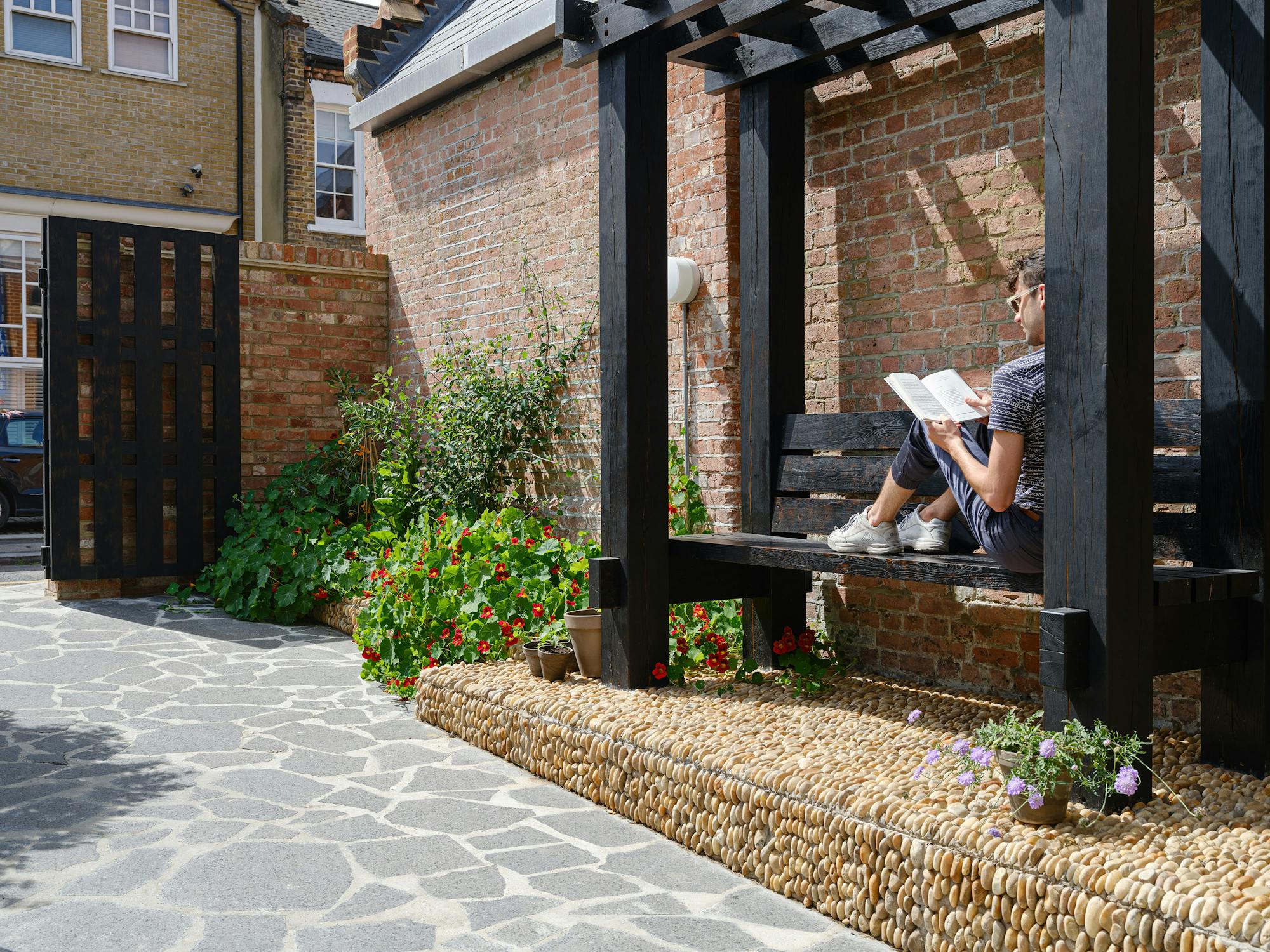 A person reads a book while sitting on a black wooden bench in a sunlit courtyard. The courtyard features a stone pathway, a pebble-lined garden bed with red and green plants, and brick walls, with a wooden pergola providing partial shade.