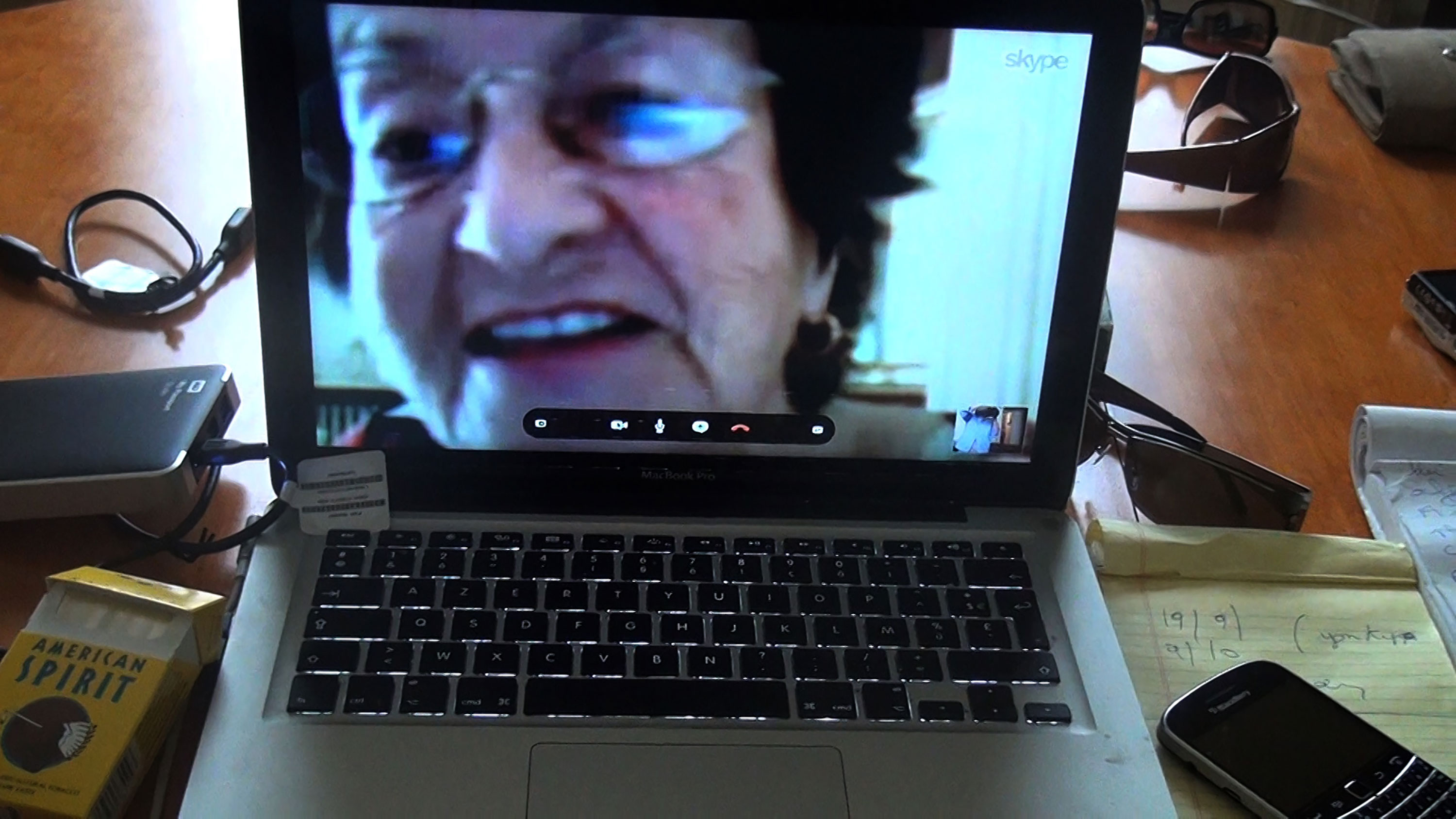 A laptop screen displays a close-up video call of an older woman with short dark hair and glasses. The laptop sits on a wooden desk cluttered with a packet of cigarettes, an external hard drive, a mobile phone, a notepad, and sunglasses.