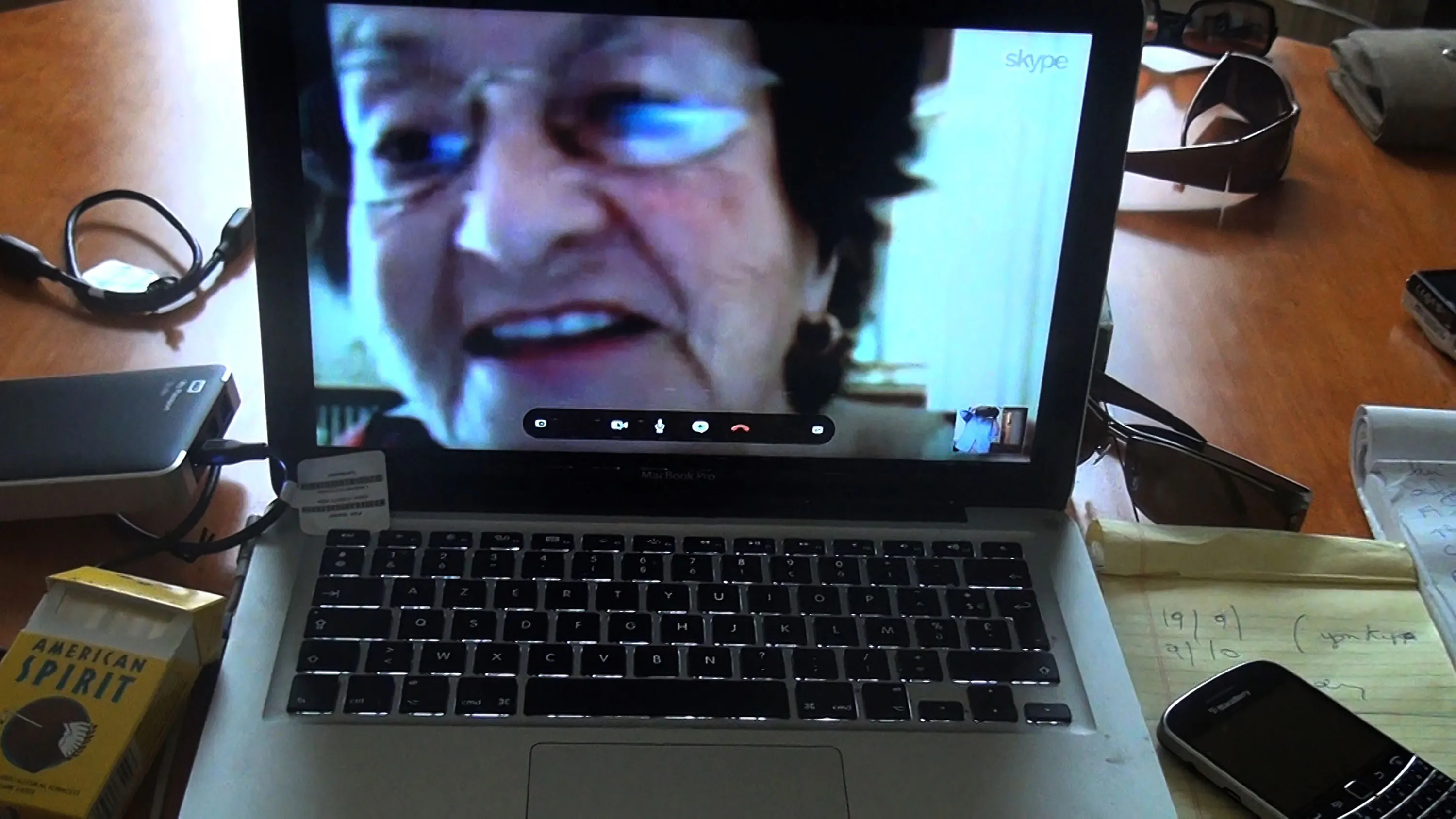 A laptop screen displays a close-up video call of an older woman with short dark hair and glasses. The laptop sits on a wooden desk cluttered with a packet of cigarettes, an external hard drive, a mobile phone, a notepad, and sunglasses.