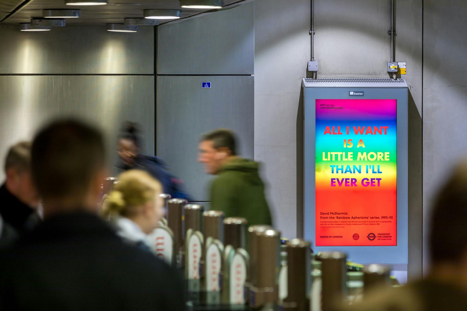 A digital billboard in a busy, modern indoor space displays colourful text: “All I want is a little more than I’ll ever get.” People walk past quickly, some blurred in motion, suggesting a rush-hour environment.