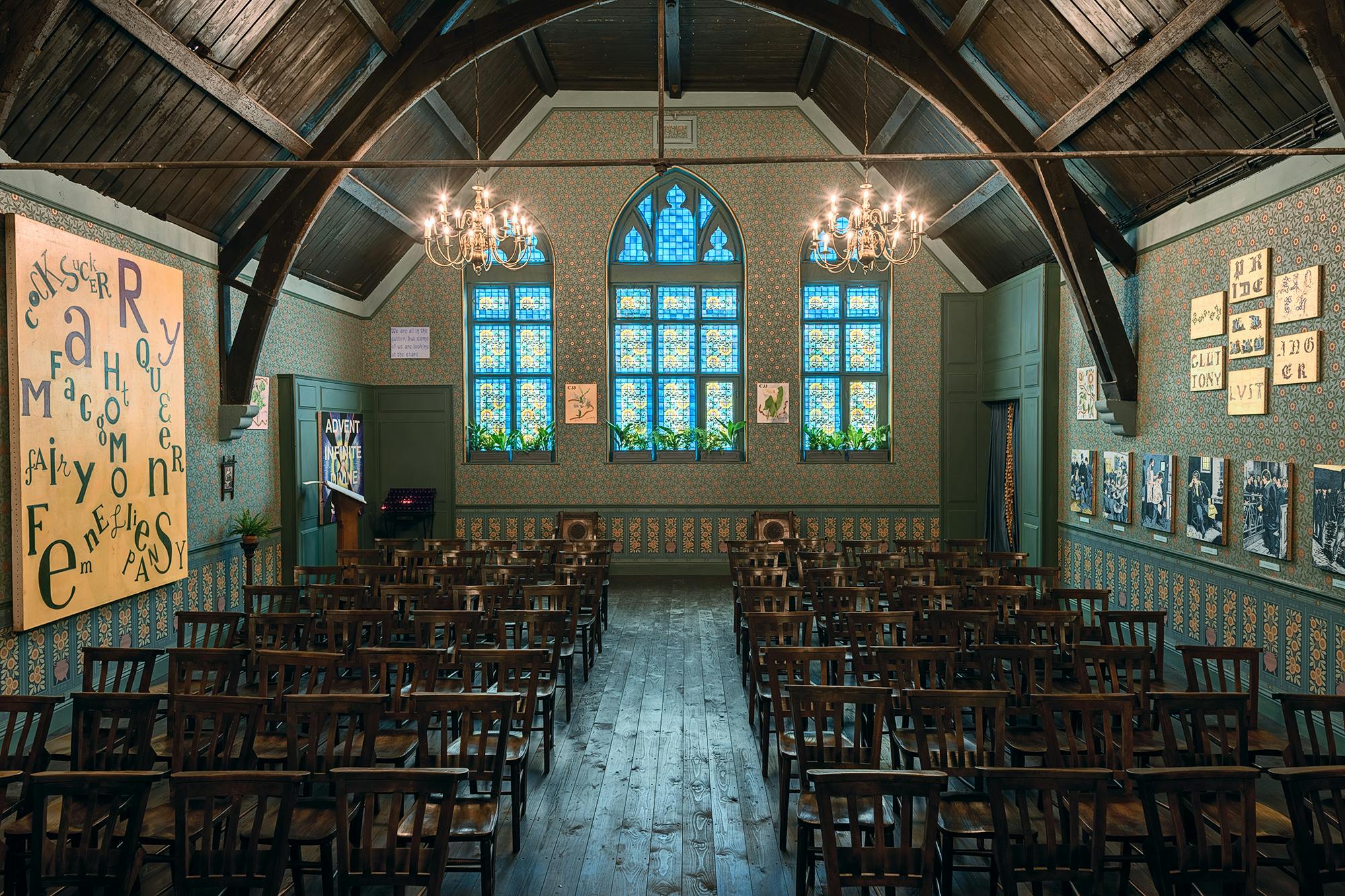 Studio Voltaire's with dark wooden floors and chairs arranged in rows facing a stage area. The walls are decorated with vintage wallpaper, framed artwork, and letter displays. A large arched window with colorful stained glass is in the centre, flanked by chandeliers.