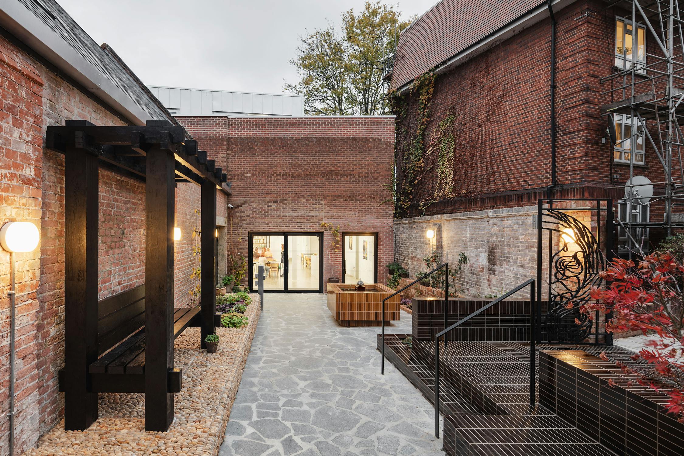 an exterior photograph of Studio Voltaire's entrance, shot from the Garden. The garden is paved with irregular shaped slabs, a raised area with steps is tiled, and a black pergola sits above a pebbled raised planted bed.