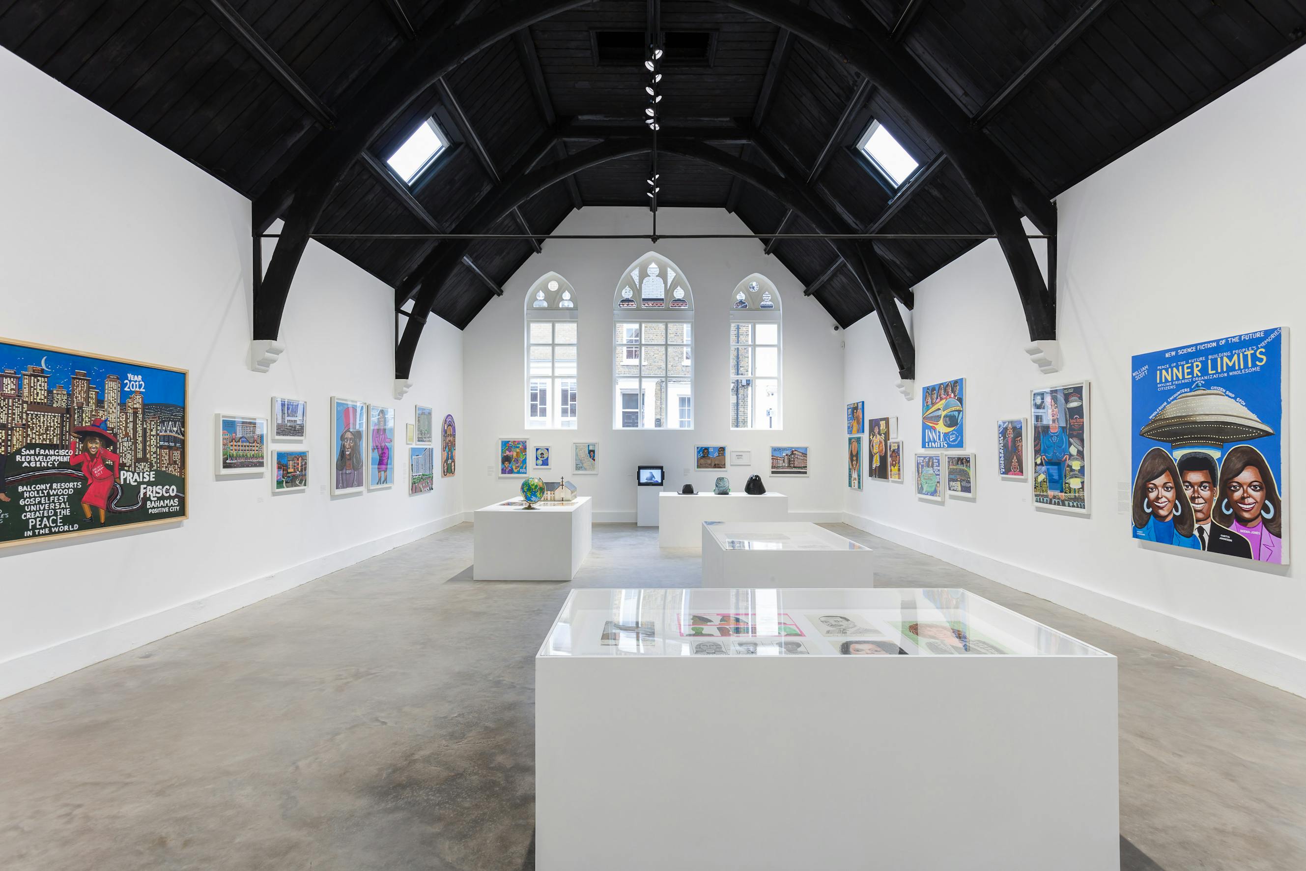 Studio Voltaire's gallery with high, dark, wooden ceilings and white walls displays colourful paintings and artworks. The space is well-lit by windows and track lighting. Several art pieces are displayed in glass cases and on walls, with a central aisle for visitors.