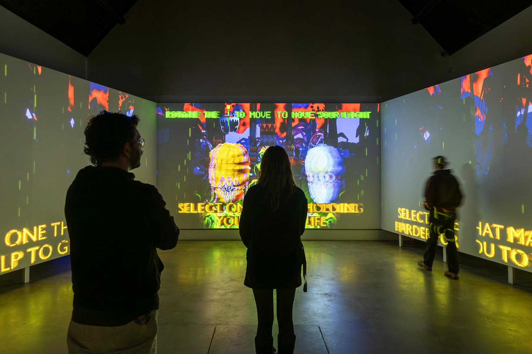 Three people  face three large wall-sized screens, projected with a game. The screen shows a landing page where users can select a playable character.