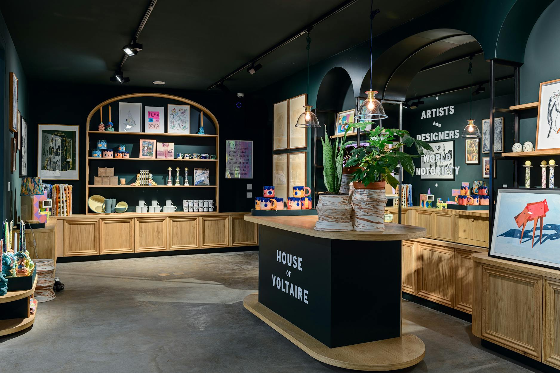 An image of House of Voltaire, a modern boutique store interior with wooden shelves displaying art prints, ceramics, and decorative objects. A counter with potted plants is in the foreground. Signs read "House of Voltaire" and "Artists, Designers, World Not Objects.
