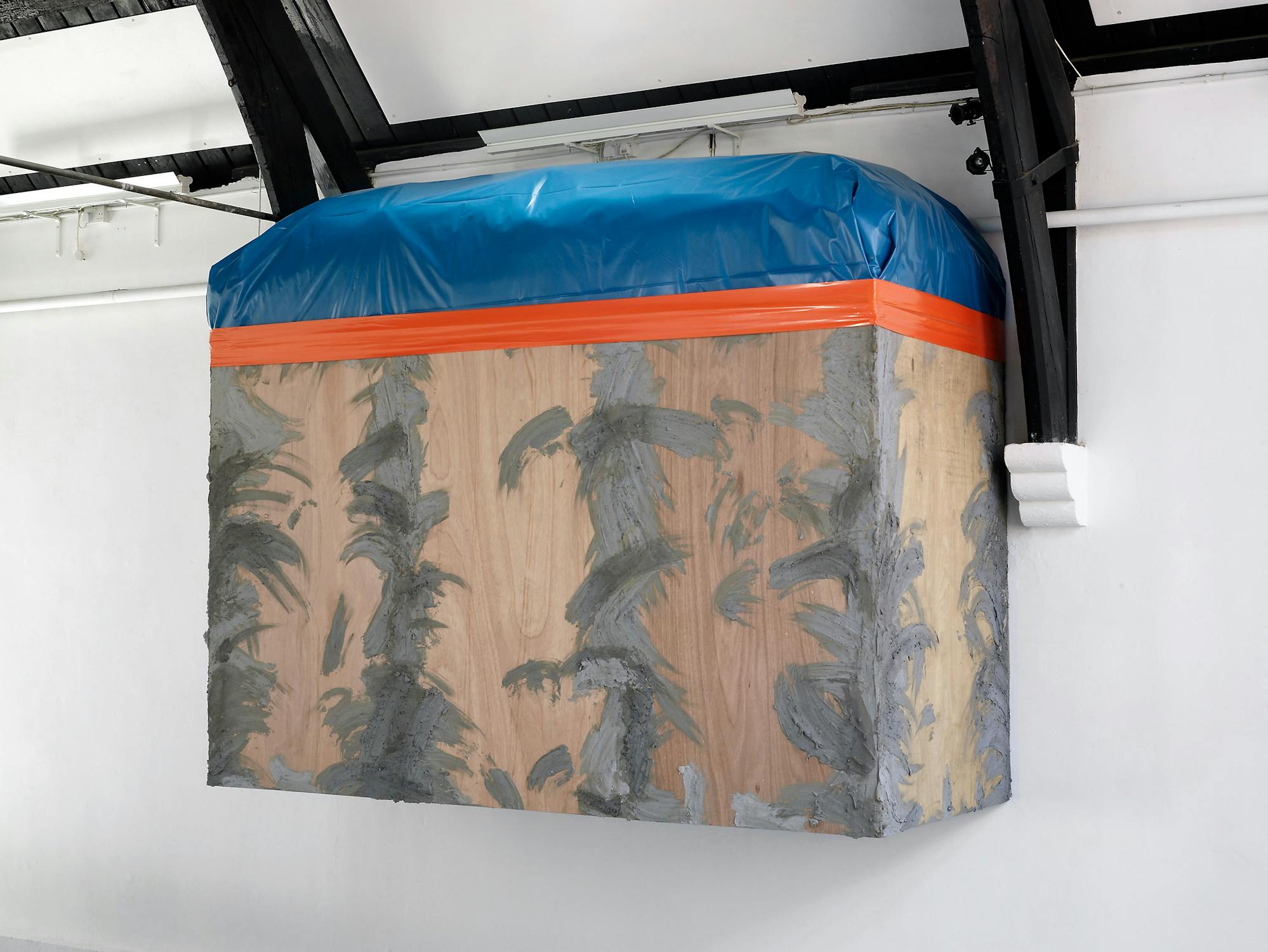 A large, rectangular wooden box is mounted on a wall near the ceiling, covered on top with a blue tarpaulin secured by an orange strap. The wood surface has rough, blotchy patches of grey material resembling cement in abstract patterns.