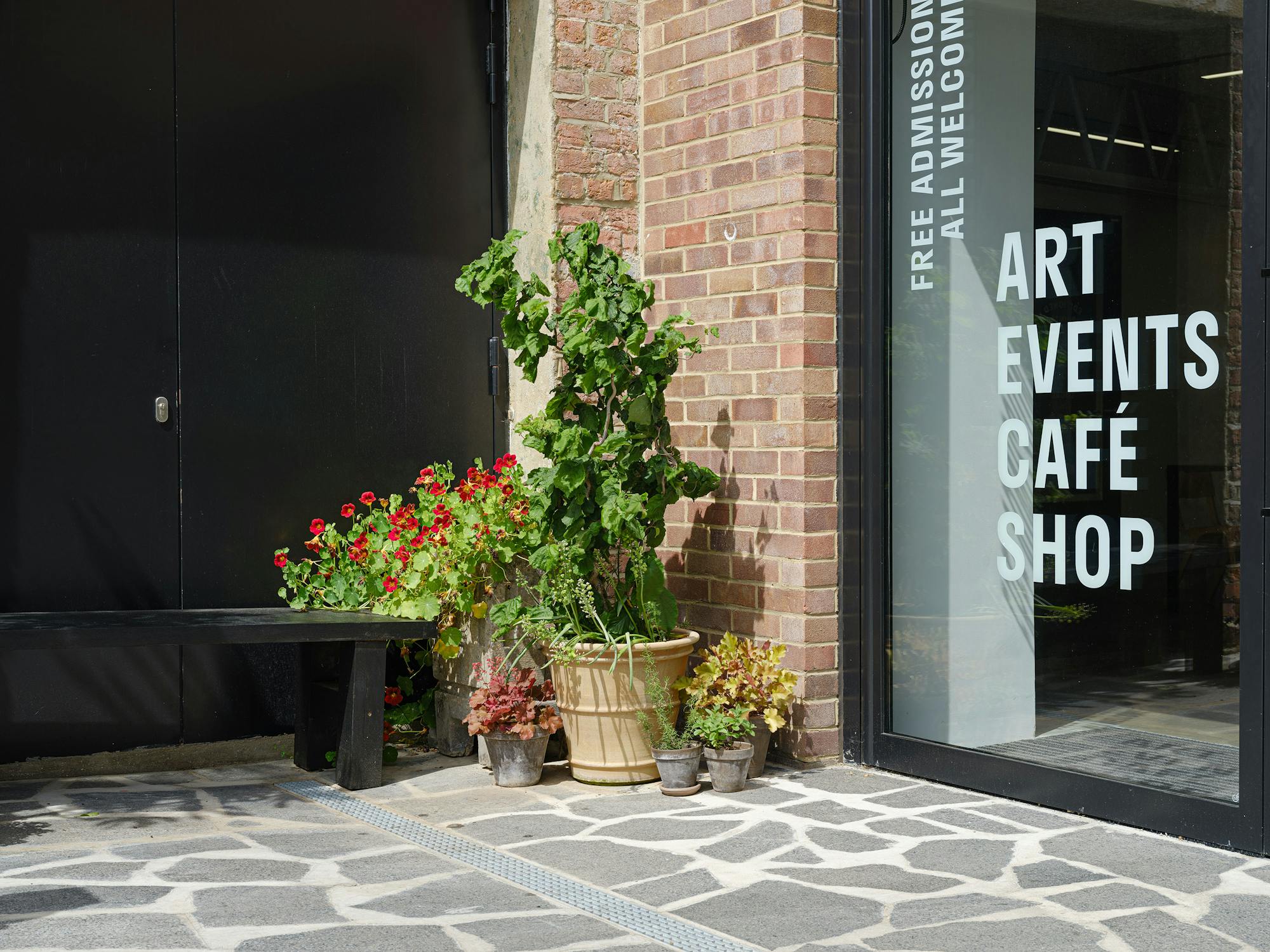 Outdoor entrance to a building with a brick exterior. A black bench is next to potted plants, including a large leafy plant and red flowers. The building's glass door has the text "Art Events Café Shop" along with "Free Admission, All Welcome."