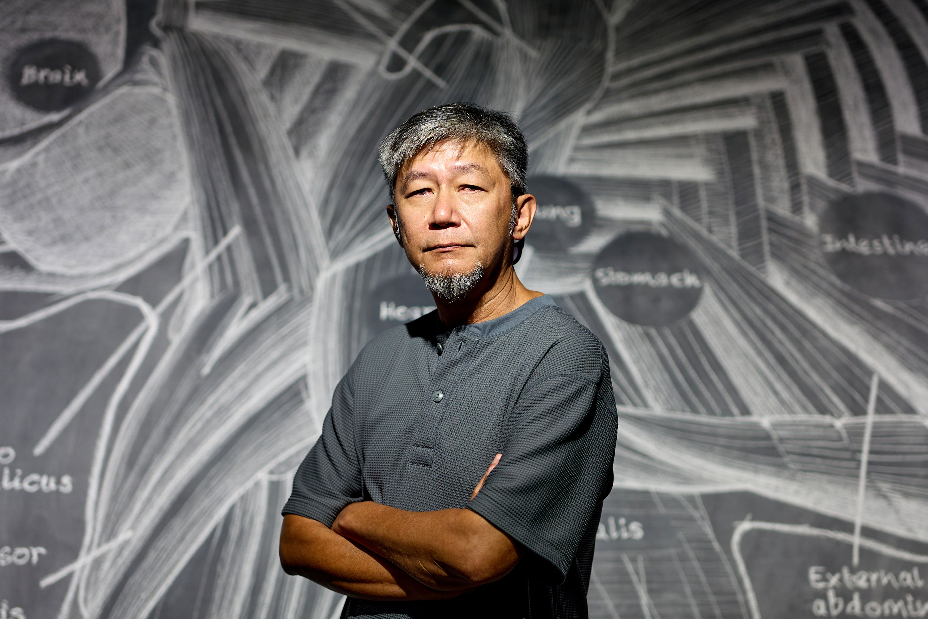Portrait of Singaporean visual artist Anthony Chin, a man with salt-and-pepper hair and a goatee, standing with folded arms and looking directly at the camera. He wears a textured grey polo shirt and stands indoors in front of a black-and-white, chalkboard-style background