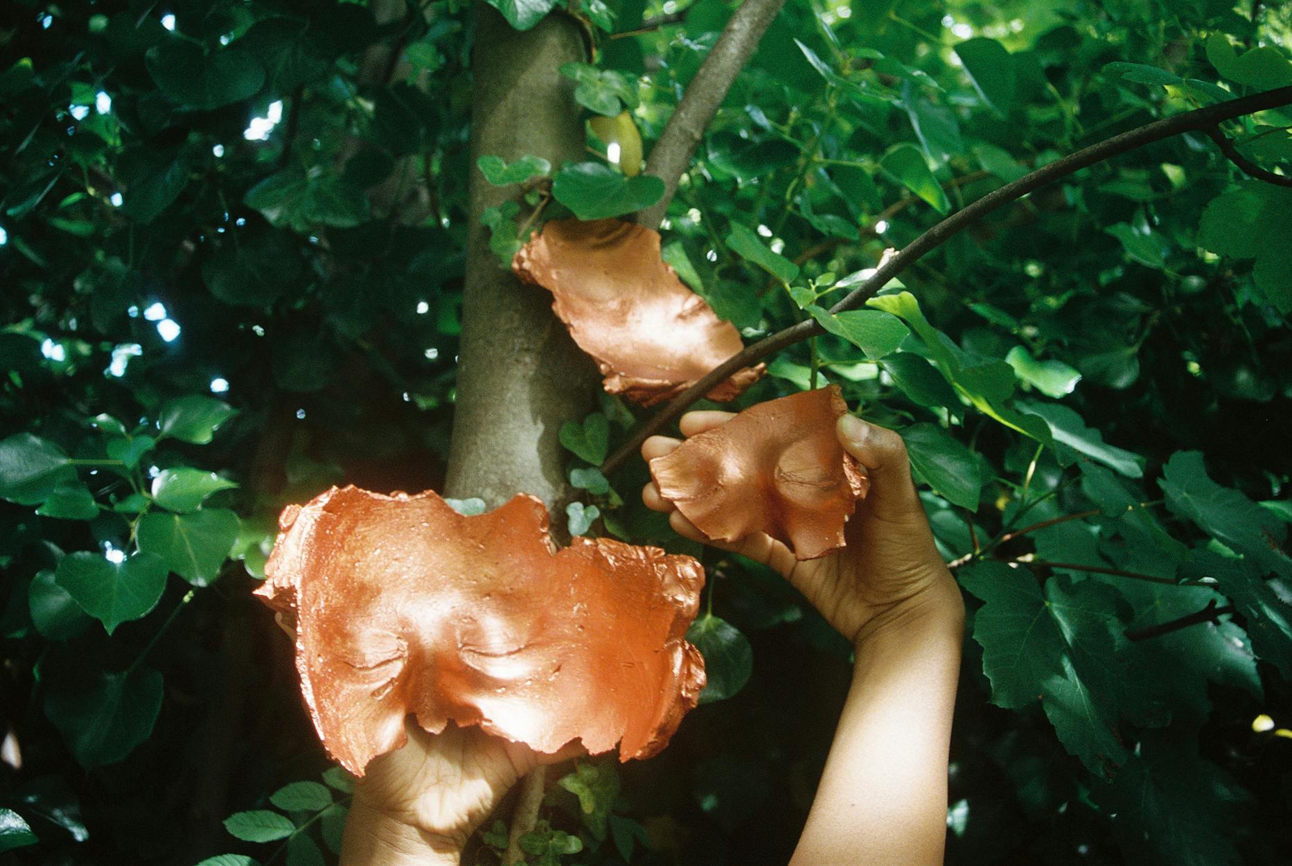 Hands holding a copper-colored mask with facial features. Similar masks are attached to the branches of a tree surrounded by green leaves, blending nature with art in a vibrant outdoor setting.