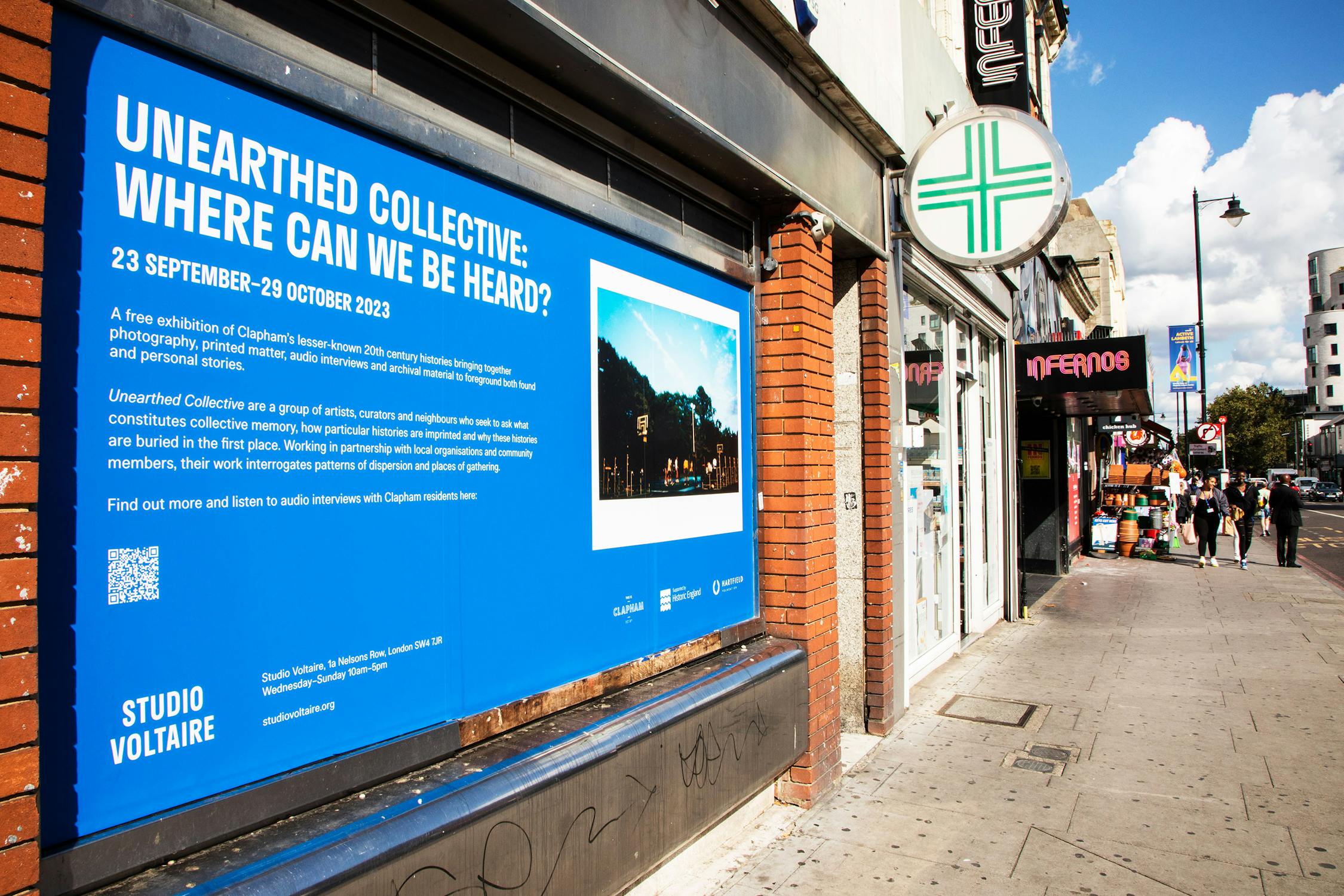 Billboard on a brick wall displaying an ad for "Unearthed Collective: Where Can We Be Heard?" with dates and location details. To the left, a poster announces another event. The scene is urban with closed shop shutters and graffiti.